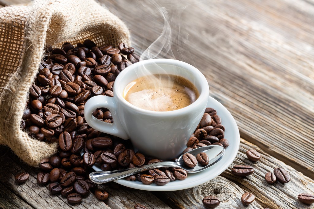 Steaming hot espresso in a white cup and saucer, surrounded by roasted coffee beans spilling from a burlap sack on a wooden table.
