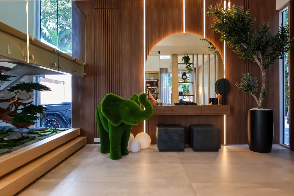 Lobby with wooden slatted walls, a large oval mirror, a topiary elephant, a potted olive tree, and a glass-railed staircase.