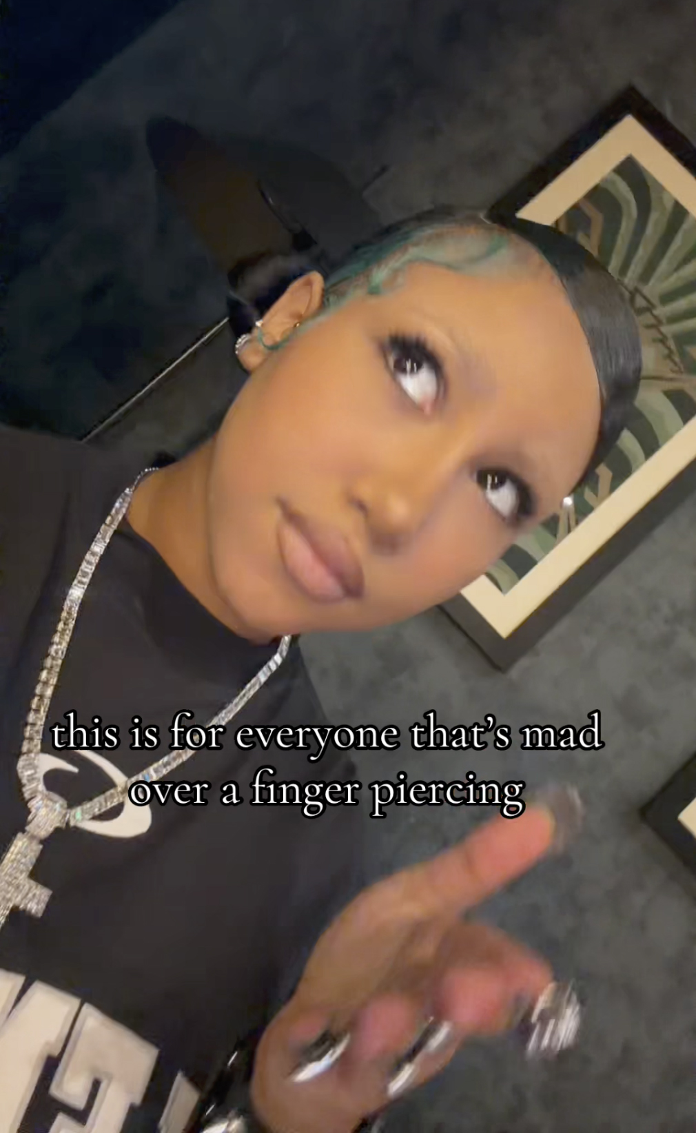 North West with blue-tinted hair and a diamond necklace holds out their hand to show off a finger piercing.