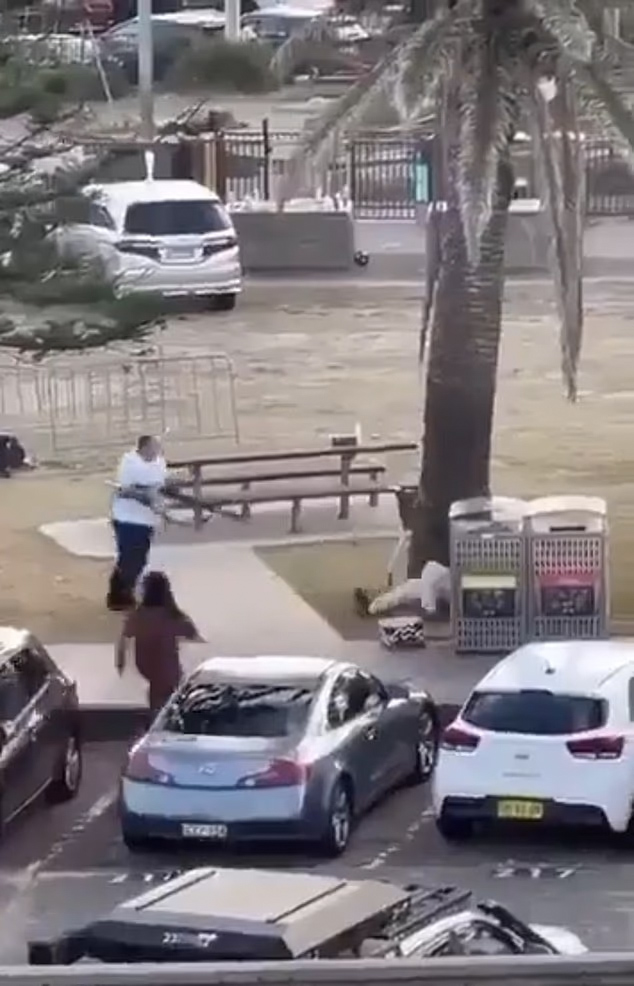 A man being assaulted by a person with a weapon while a third person runs away from the scene, with cars and a palm tree in the foreground.