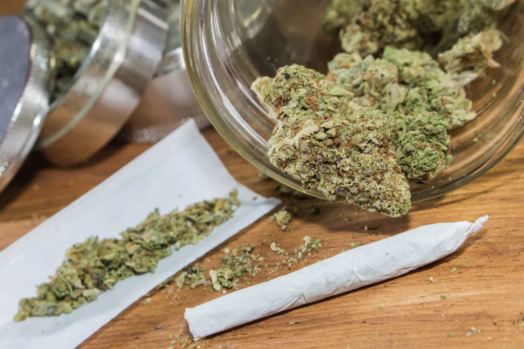 Cannabis buds in a glass jar, alongside a rolling paper with ground cannabis, and a rolled joint on a wooden surface.