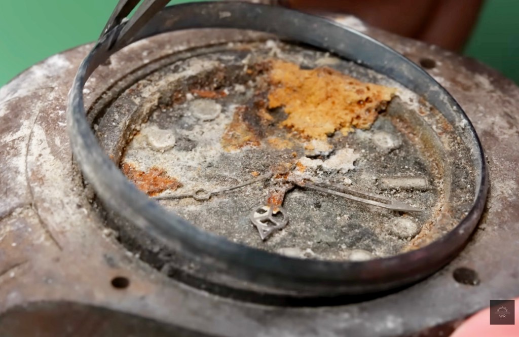 A rusty, corroded watch face with hands frozen in place, surrounded by a metal ring held by tweezers.