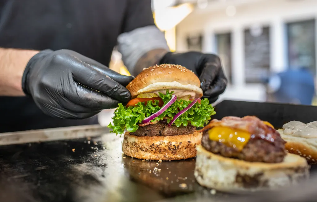 A person wearing black gloves preparing a fresh burger on a grill with another burger with melted cheese and bacon in the foreground.