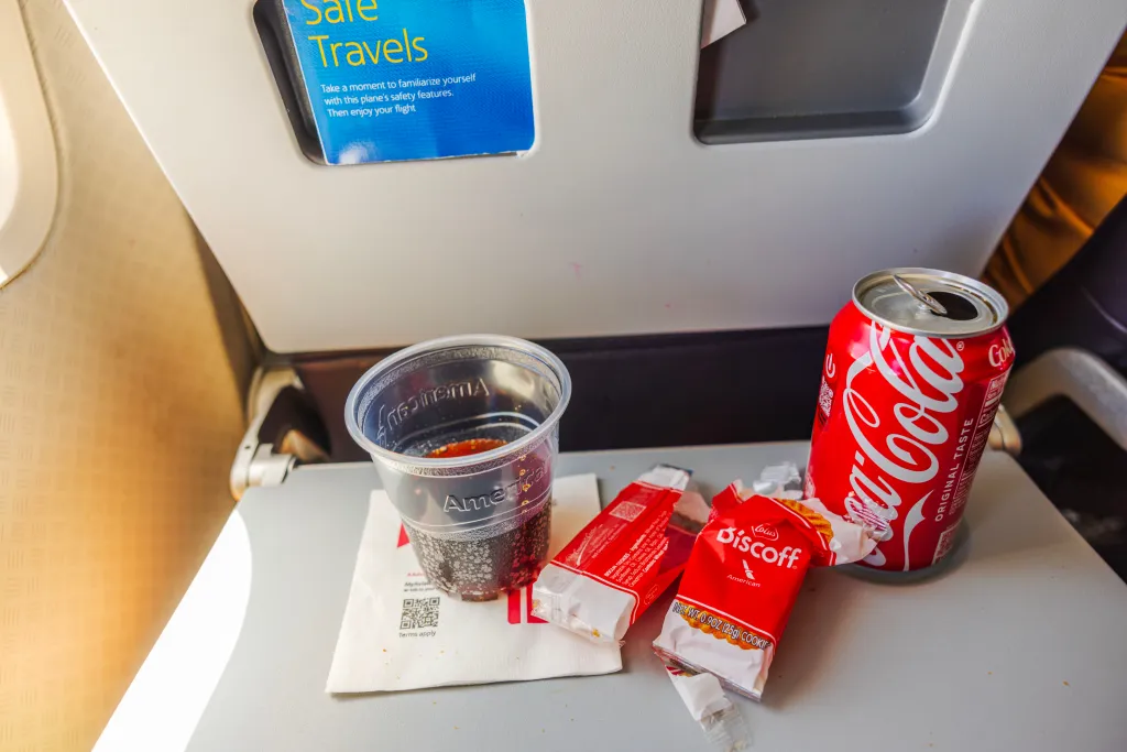 Finished American Airlines in-flight breakfast with an empty Coca-Cola can, a partially full cup of Coca-Cola, and empty Biscoff cookie wrappers on a tray table.