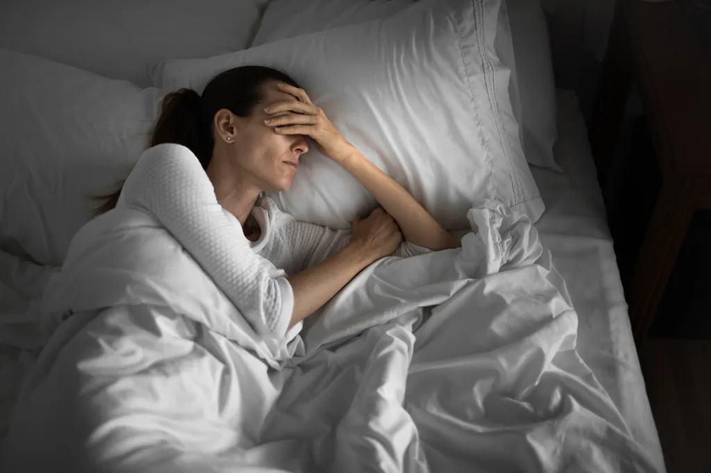 A woman lies in bed, one hand covering her eyes, appearing troubled or in pain.