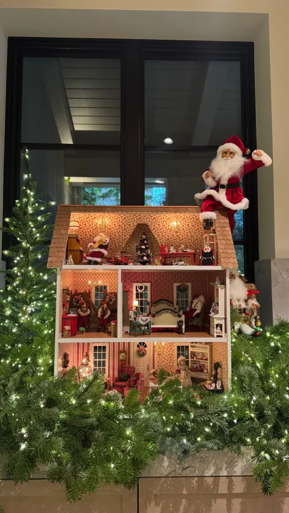 A Christmas-themed dollhouse with miniature Santa Claus figures and lit evergreen garlands.