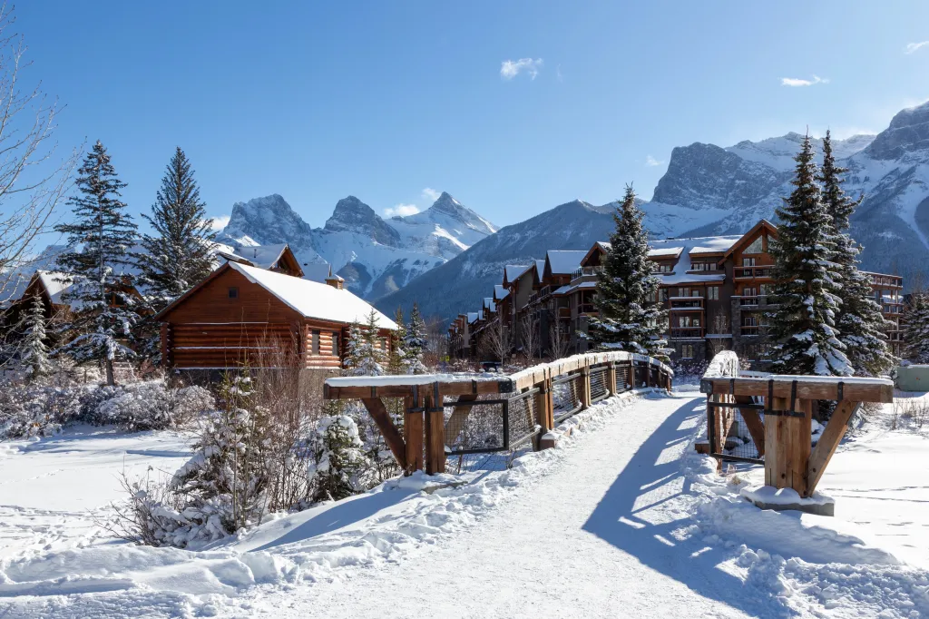 Wooden pedestrian footbridge in Spring Creek Mountain Village, Canmore, Alberta, with the snow-covered Three Sisters mountains in the distance.