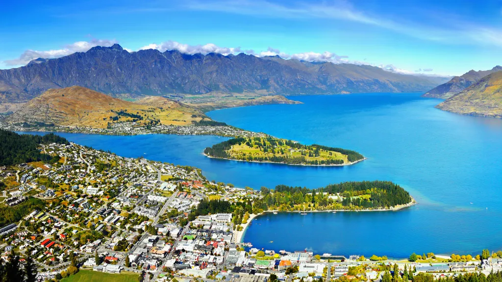 Aerial view of Queenstown, New Zealand, with a town along the blue lake and mountains in the background.
