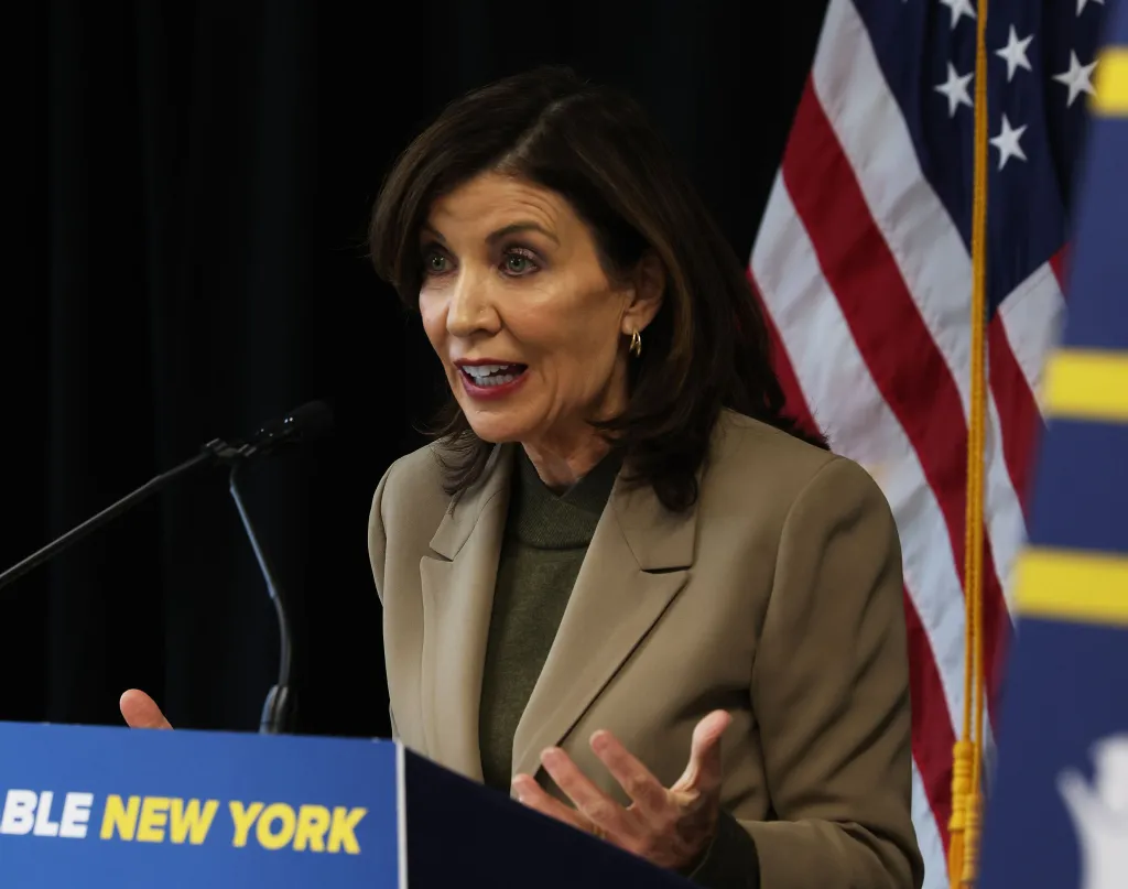 NY Governor Kathy Hochul speaking at a podium with an American flag in the background.