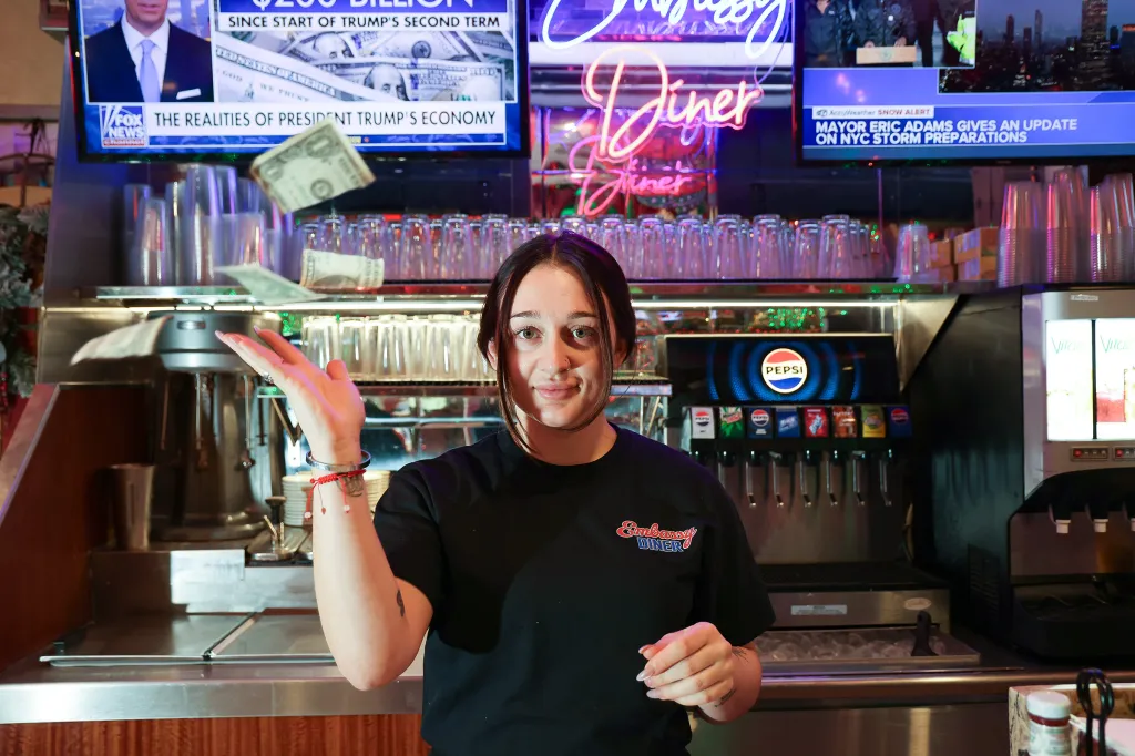 Zoe Kalodimos, a waitress at Embassy Diner, looks upset as money is thrown around, with news screens in the background.