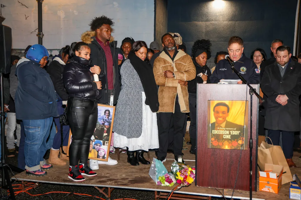A candle lighting vigil for Edeedson Cine, with people gathered on a stage and a man speaking at a podium with a photo of Edeedson Cine on it.