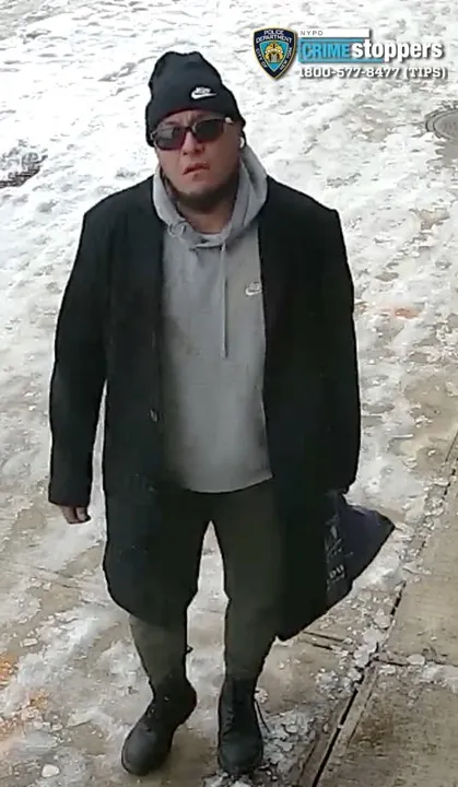 A man in a black beanie, sunglasses, gray hoodie, black coat, and dark pants carrying a dark bag, walking on a partially snow-covered sidewalk.
