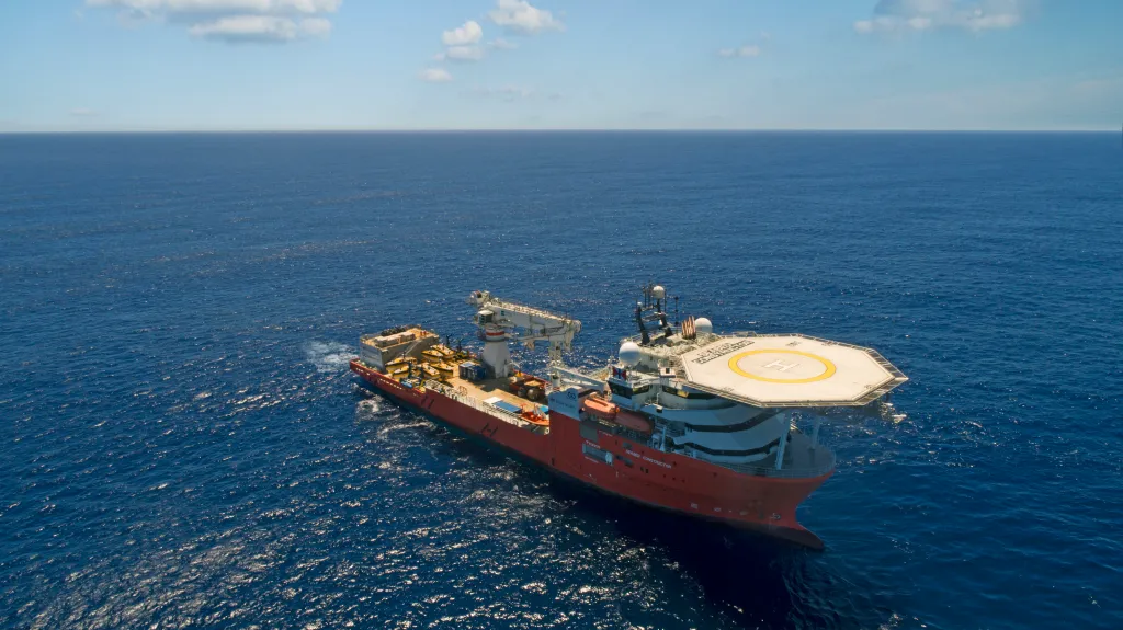 A large red ship with a helipad on its stern in the middle of the deep blue ocean, on a clear day with scattered clouds.