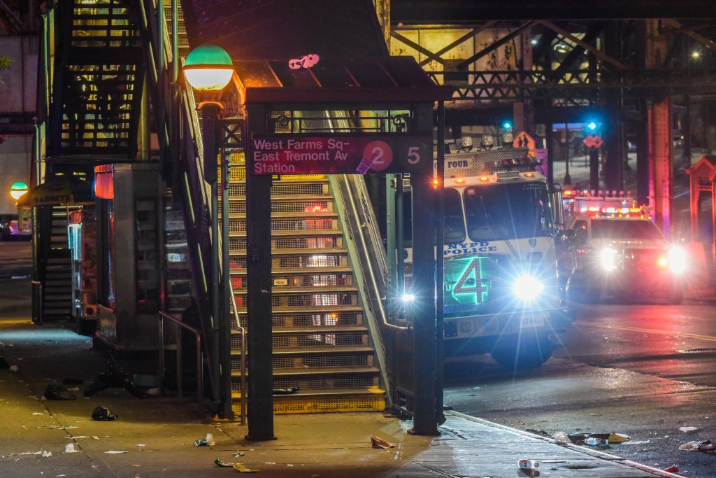 The entrance to West Farms Square-East Tremont Avenue train station at night, with an NYPD police truck and other emergency vehicles with flashing lights on the street in the background.