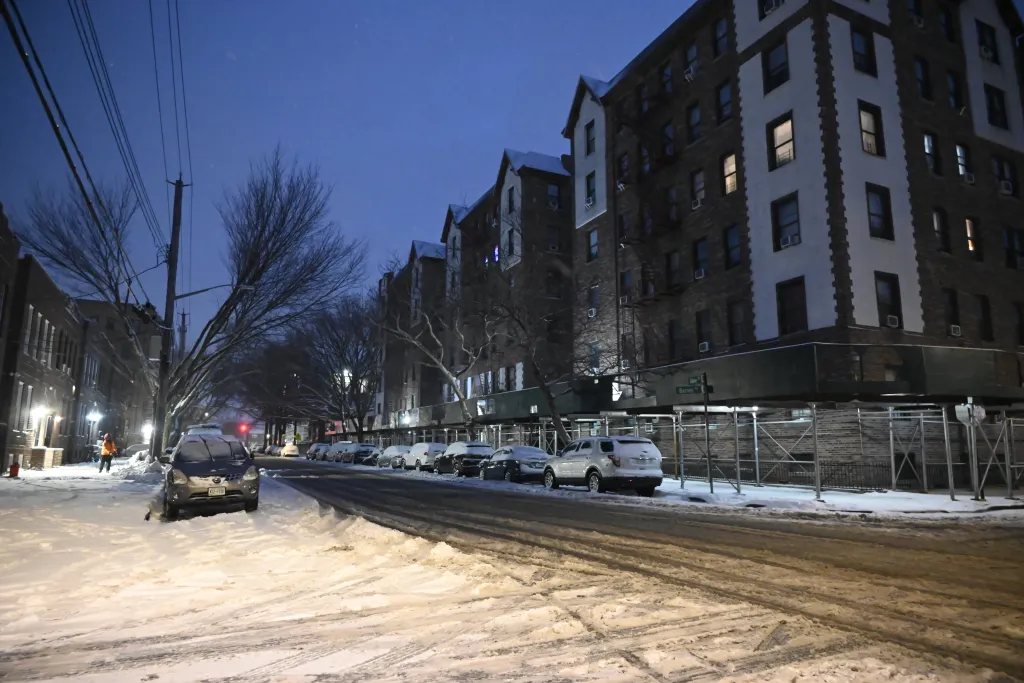 A snowy street scene with cars parked on both sides, apartment buildings, and a person shoveling snow.