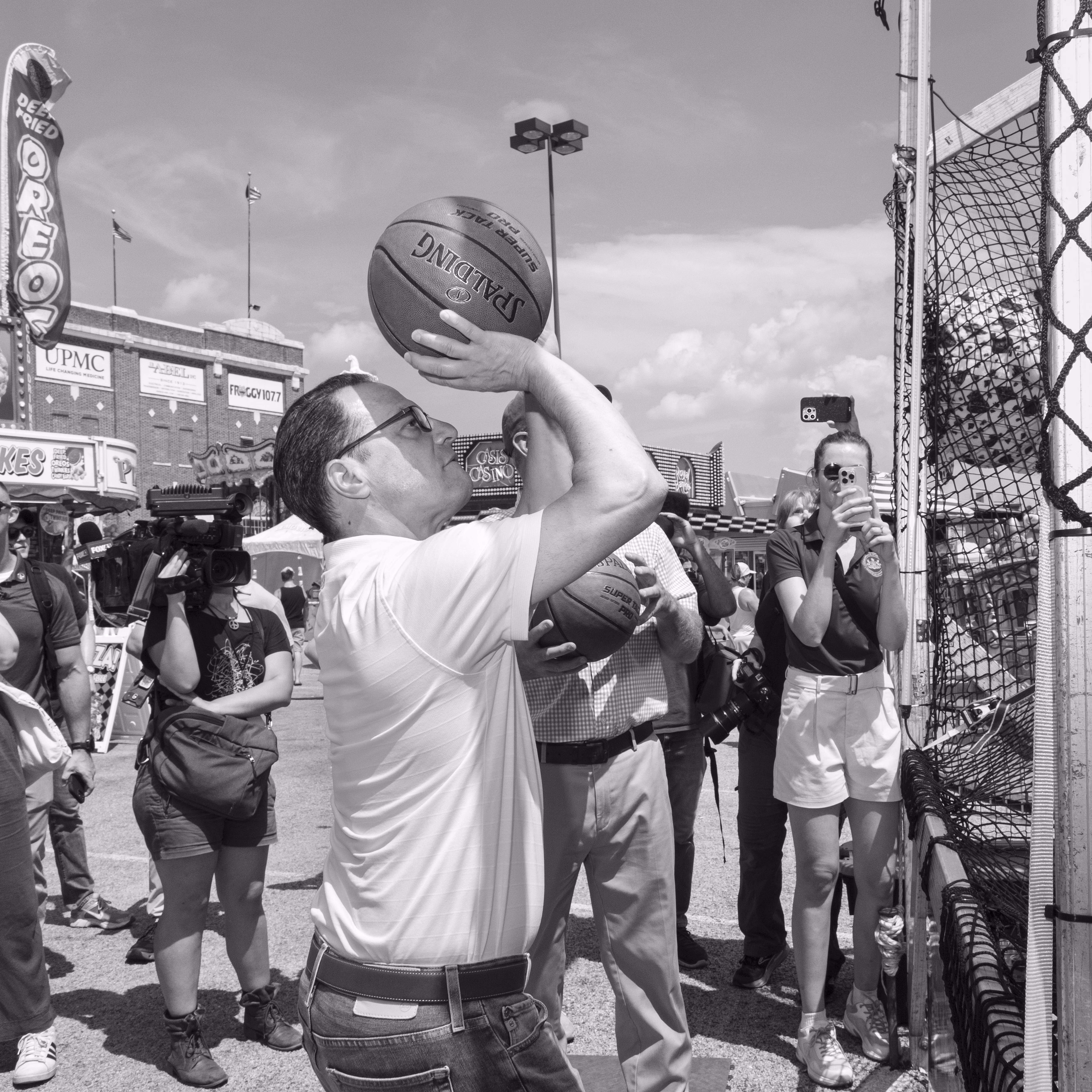 A black and white photograph of Josh Shapiro shooting a basketball into a net at a concession stand at the State Fair