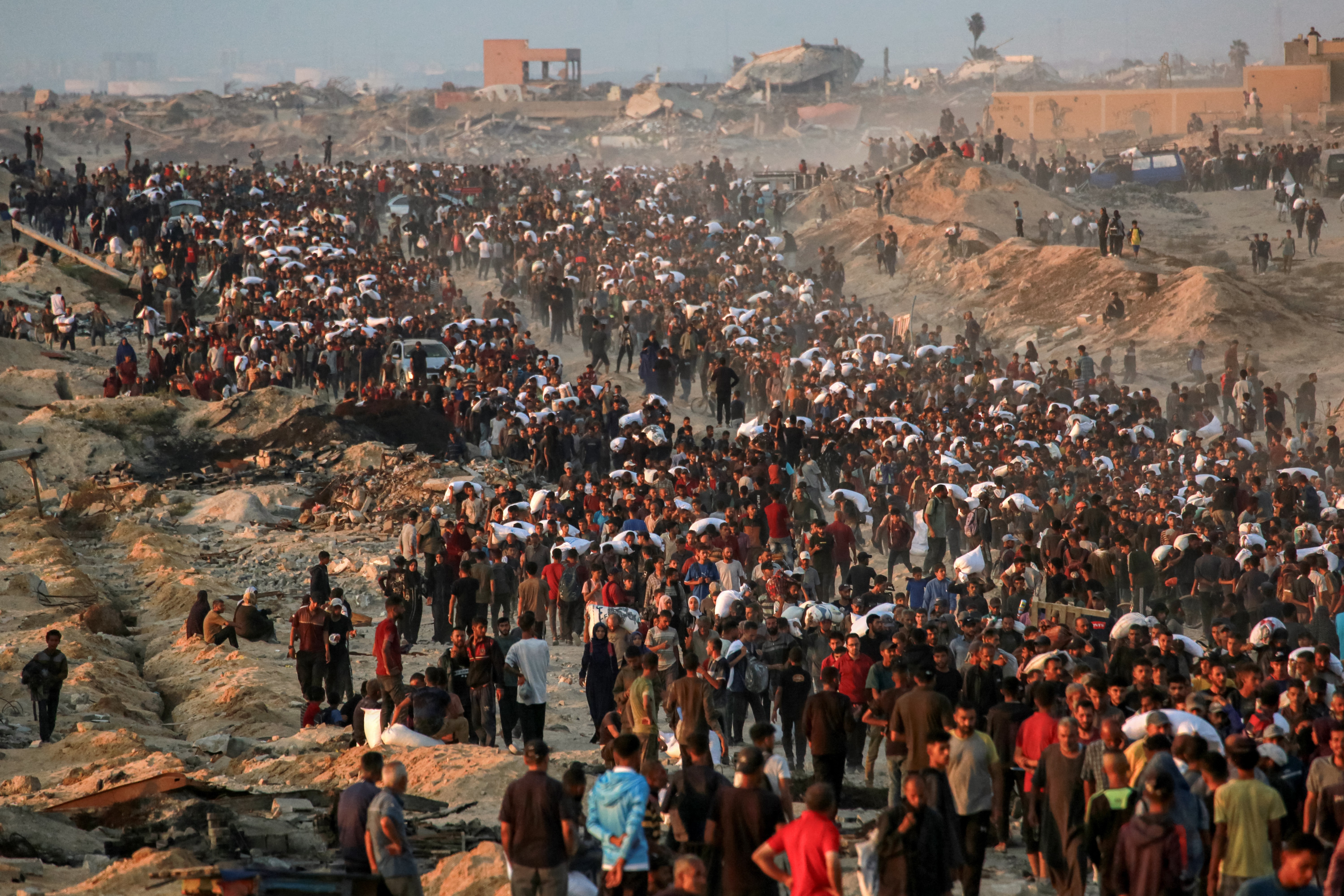 A large crowd of Palestinians carry sacks of flour, walking along a dirt road past destroyed buildings.