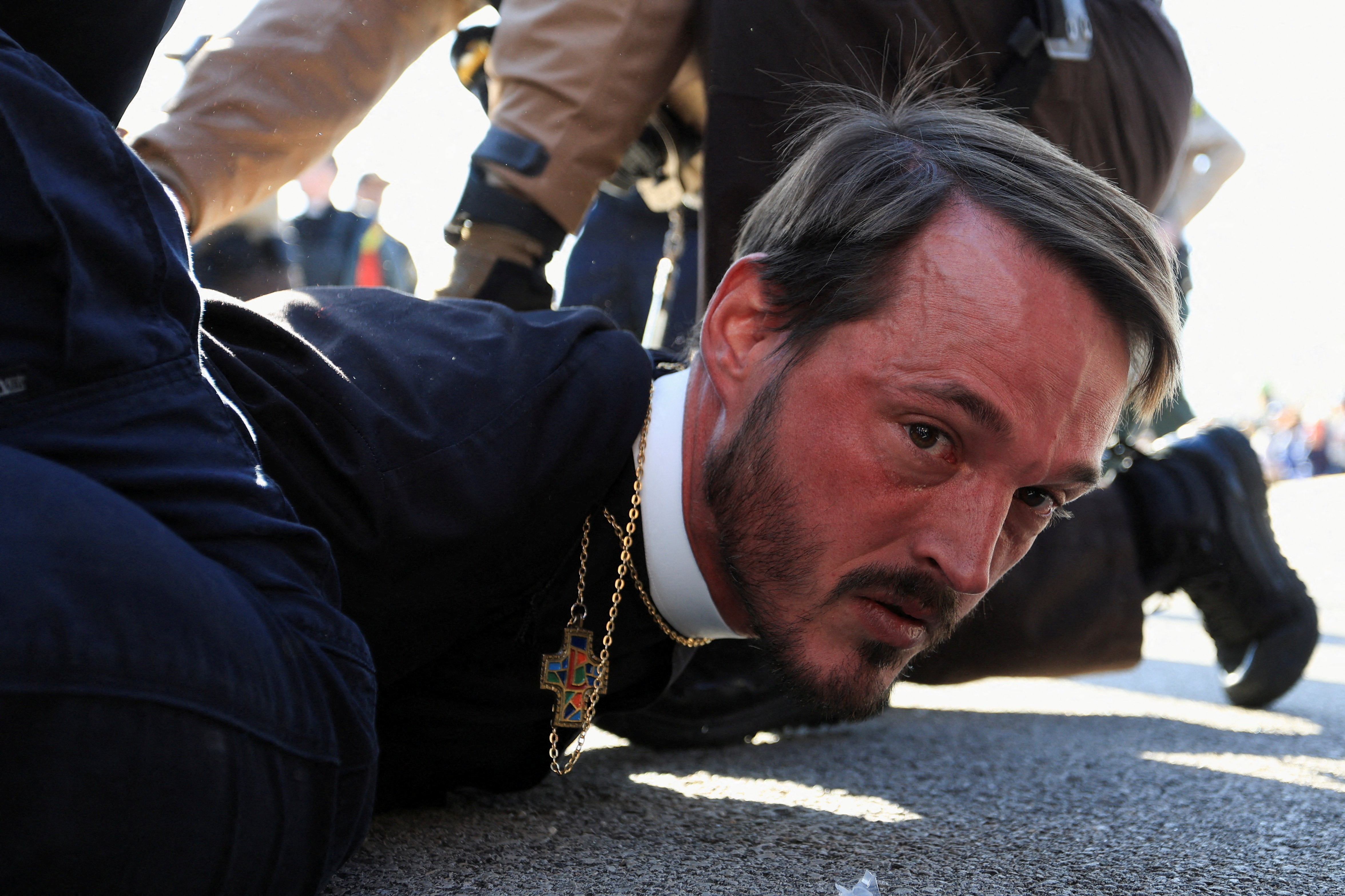 A clergyman looks toward the camera as he lies on his stomach, being detained by police officers during a protest.
