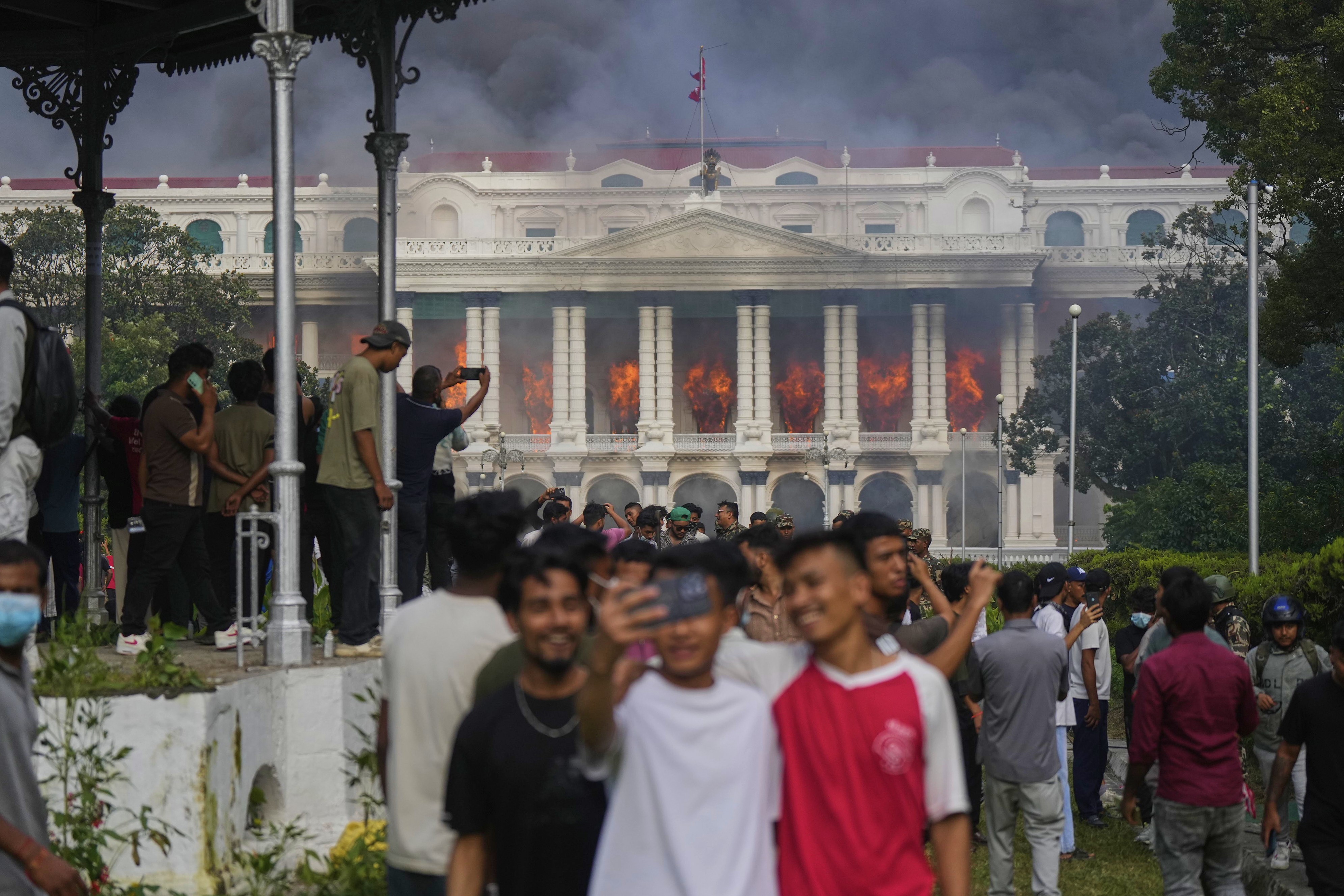 Many demonstrators mill about, while some take selfies, in front of a burning government building.