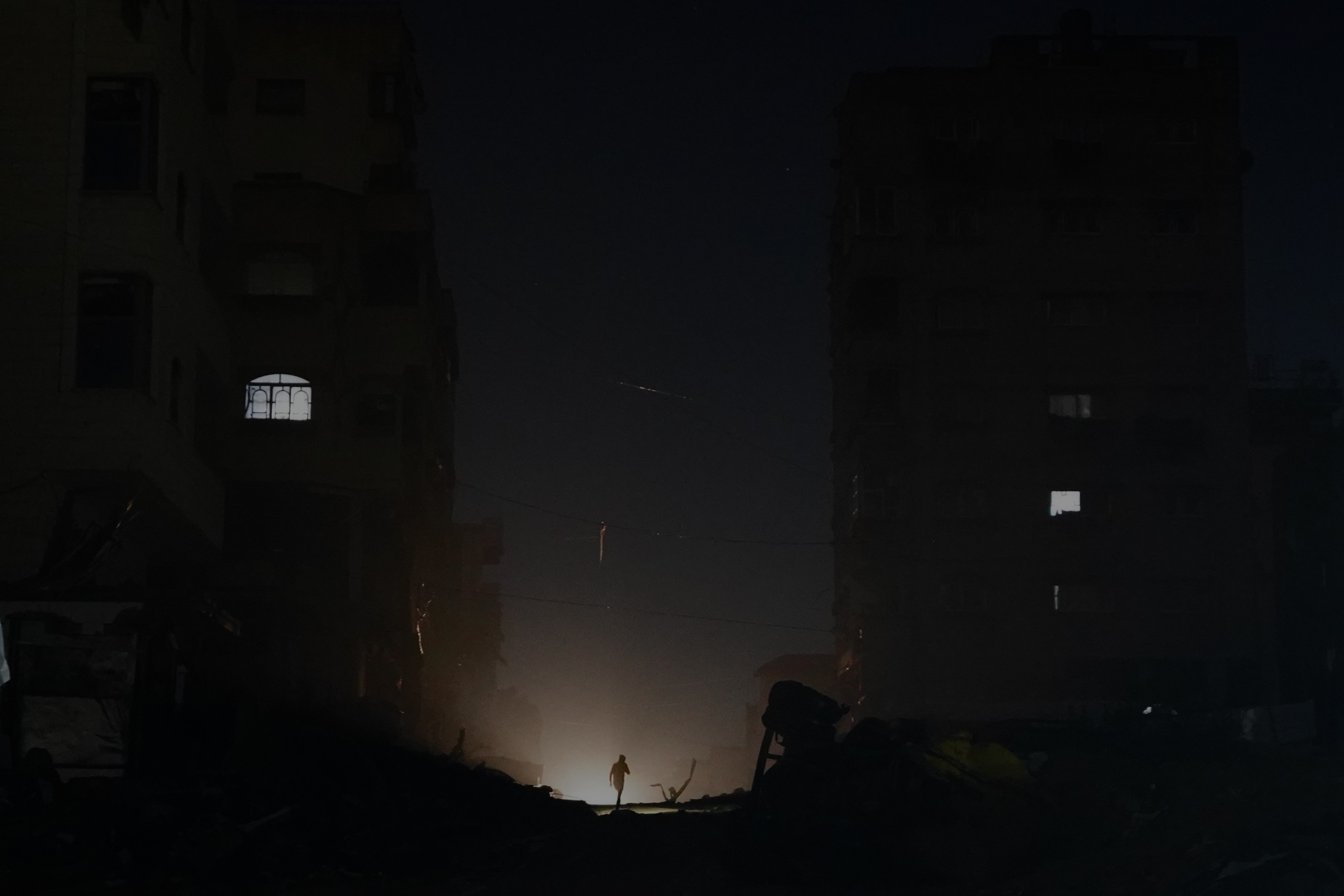 A person walks among collapsed buildings at night, silhouetted against a distant light.