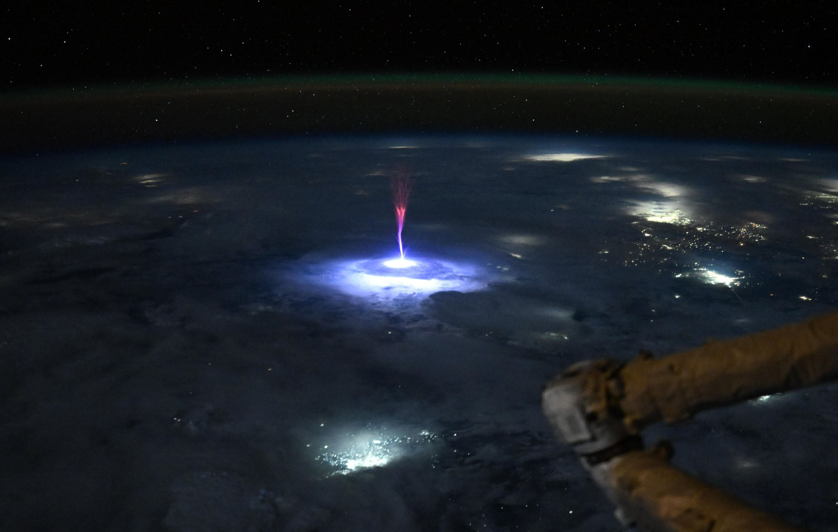 A nighttime view of the Earth, from orbit, with a bright blu-and-red bolt of electrical energy shooting up from a lightning strike in a storm below
