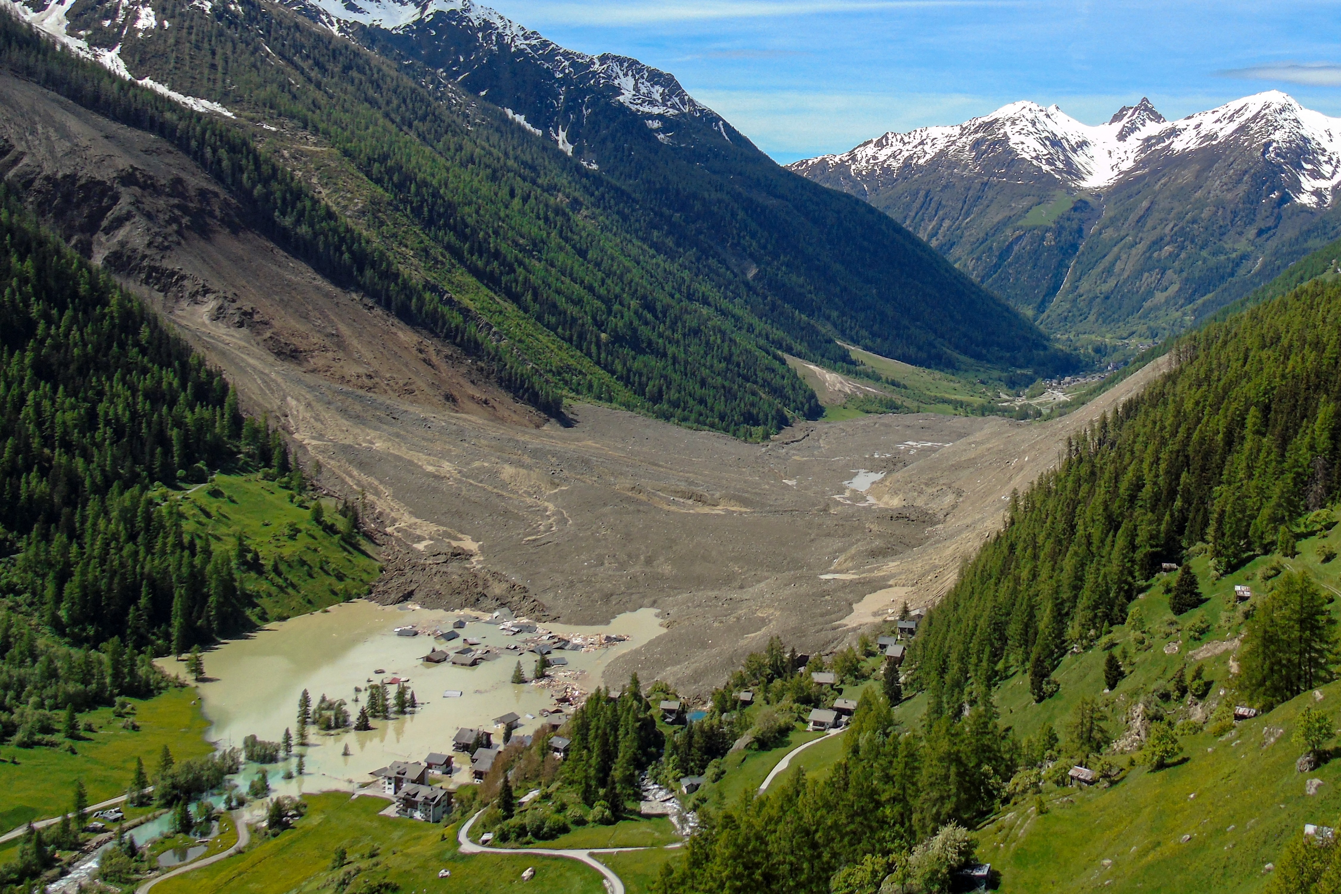 An aerial view of an Alpine valley after a massive landslide, with a cluster of houses being flooded on the uphill side
