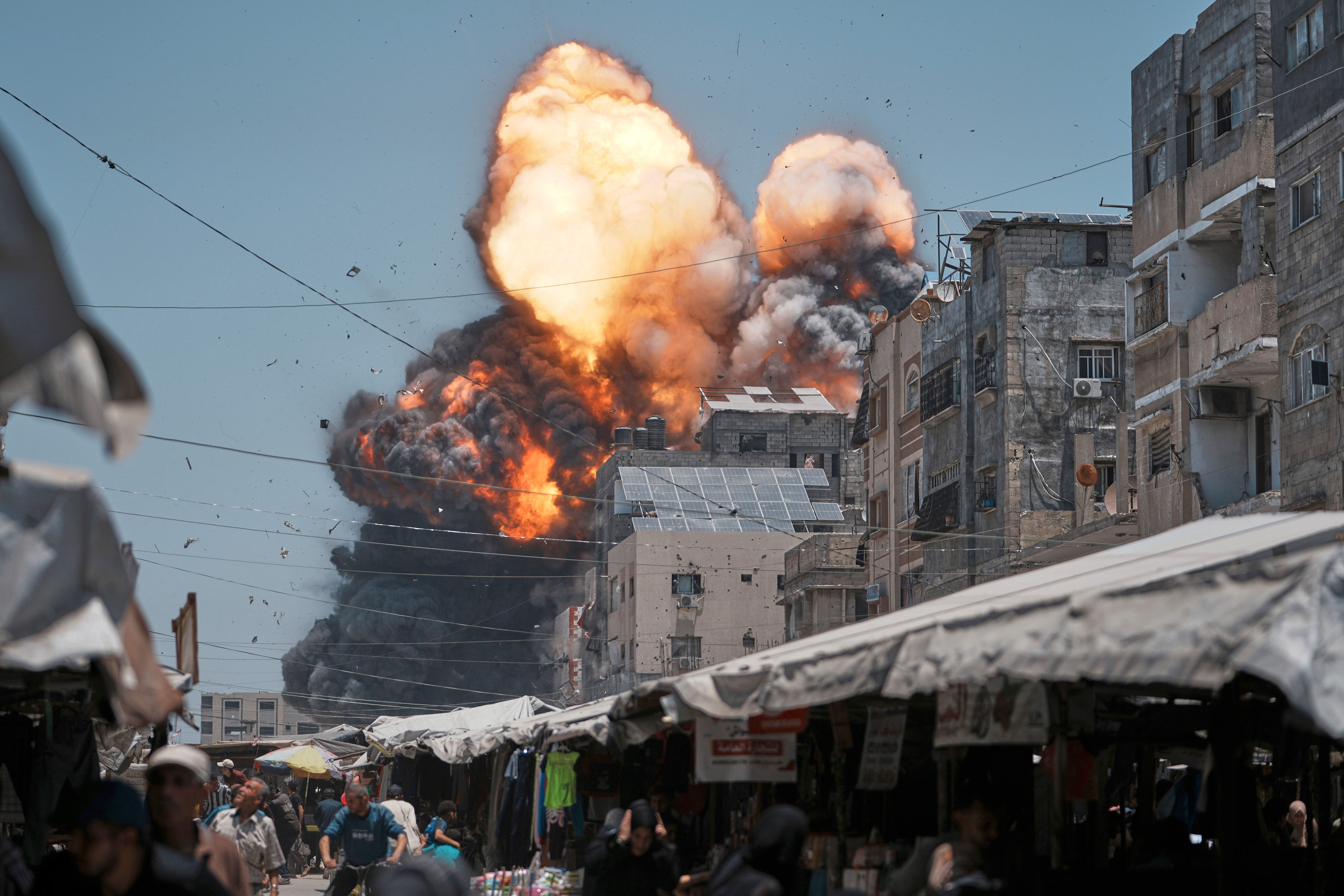 Thick smoke and flames erupt from an Israeli airstrike, as civilians run in a market the foreground.