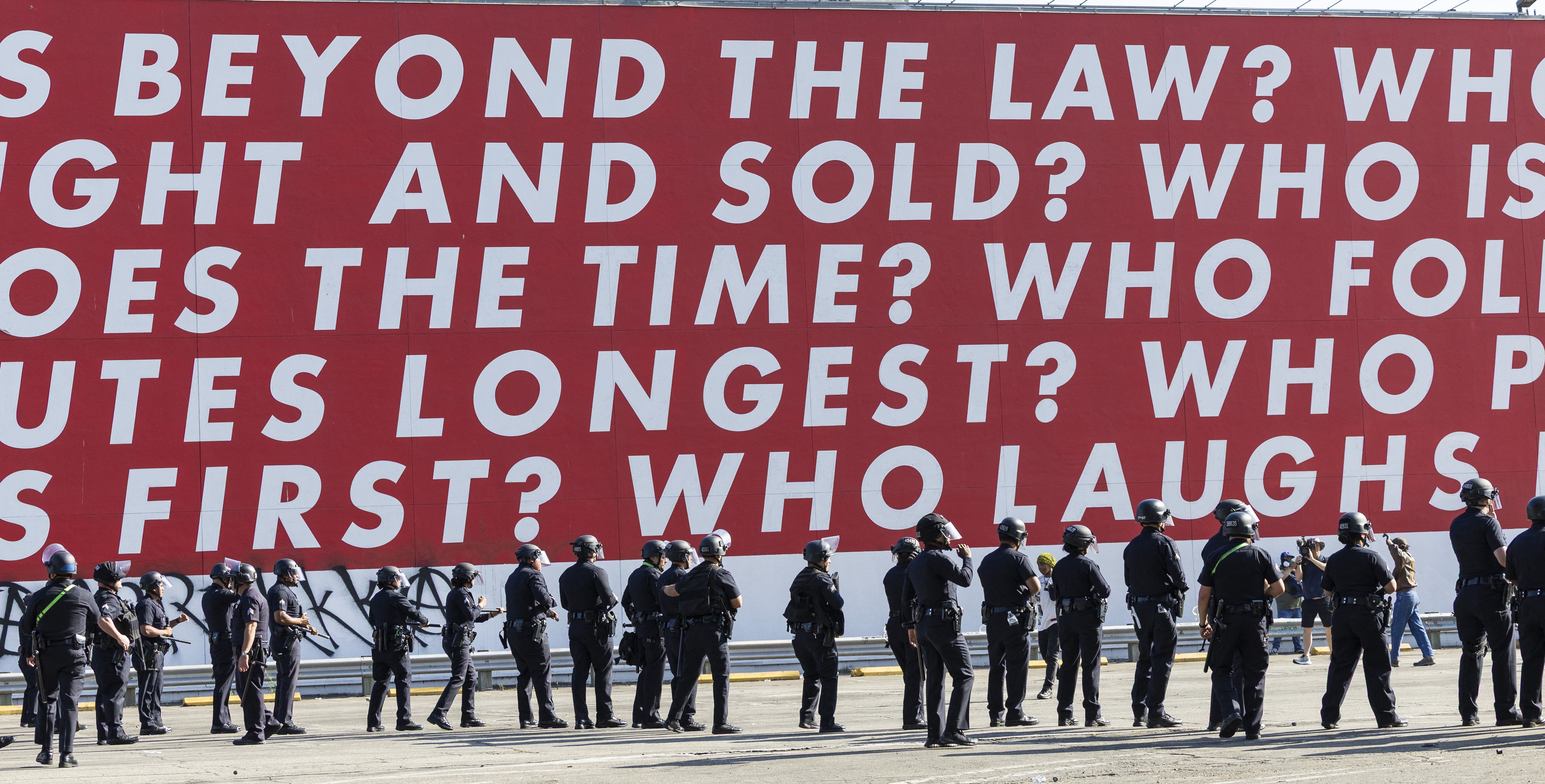 A line of riot police officers stand beneath a huge work of art on an outside wall, made up of text, reading in part 'beyond the law?'