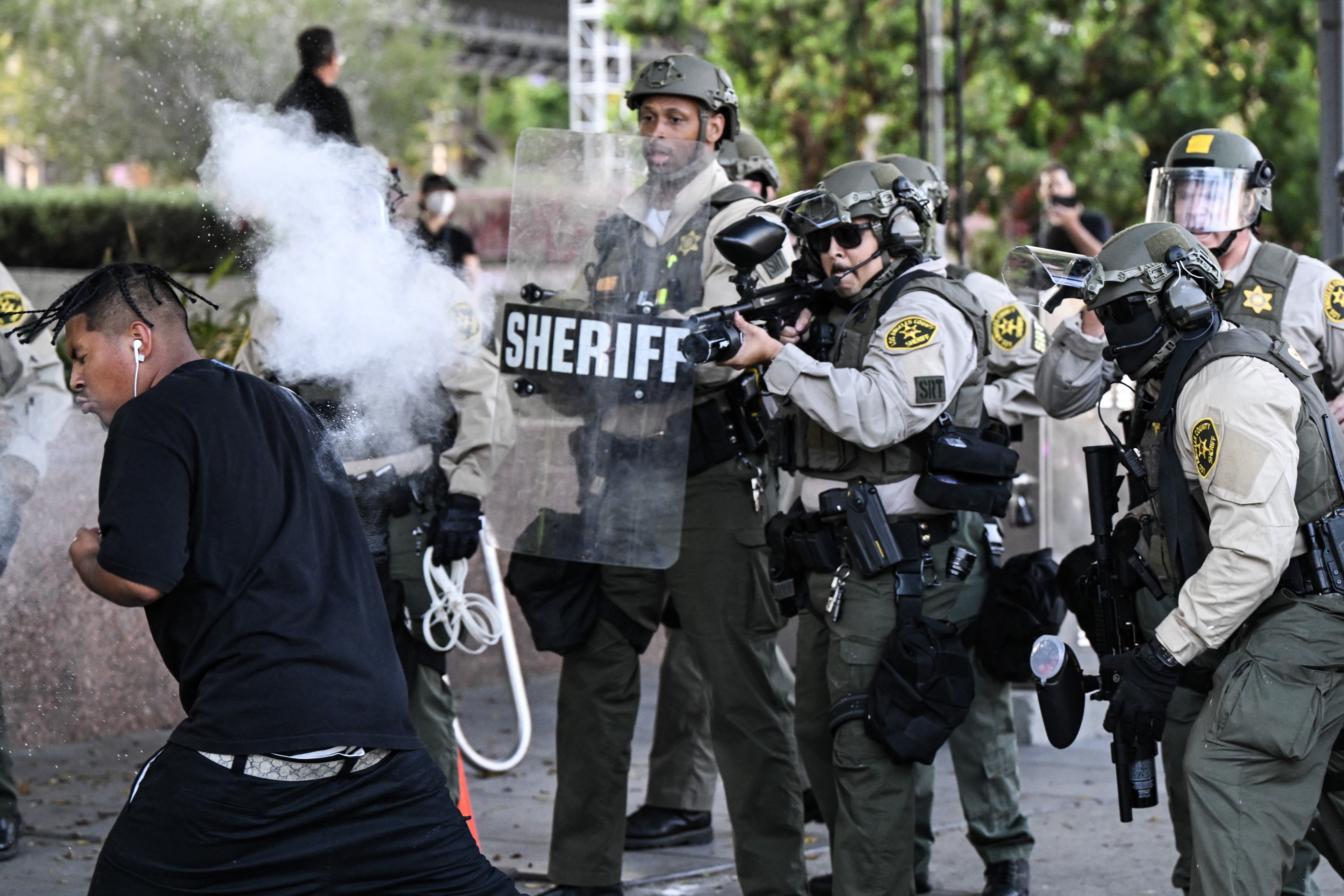 Several riot police officers face off against protesters. One officer fires a nonlethal weapon at a protester at close range.