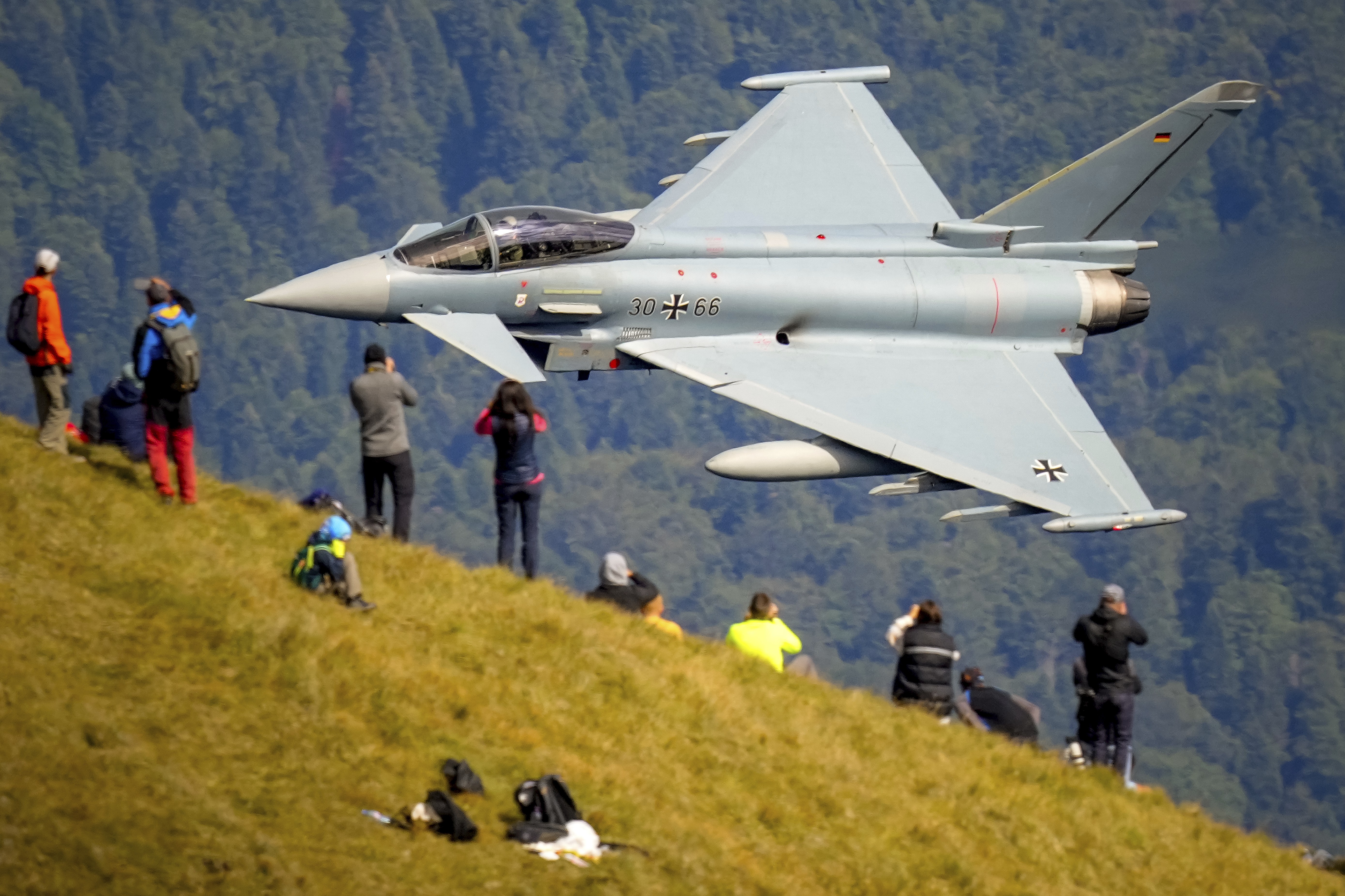 People stand on a hillside watching as a jet fighter flies past very closely.