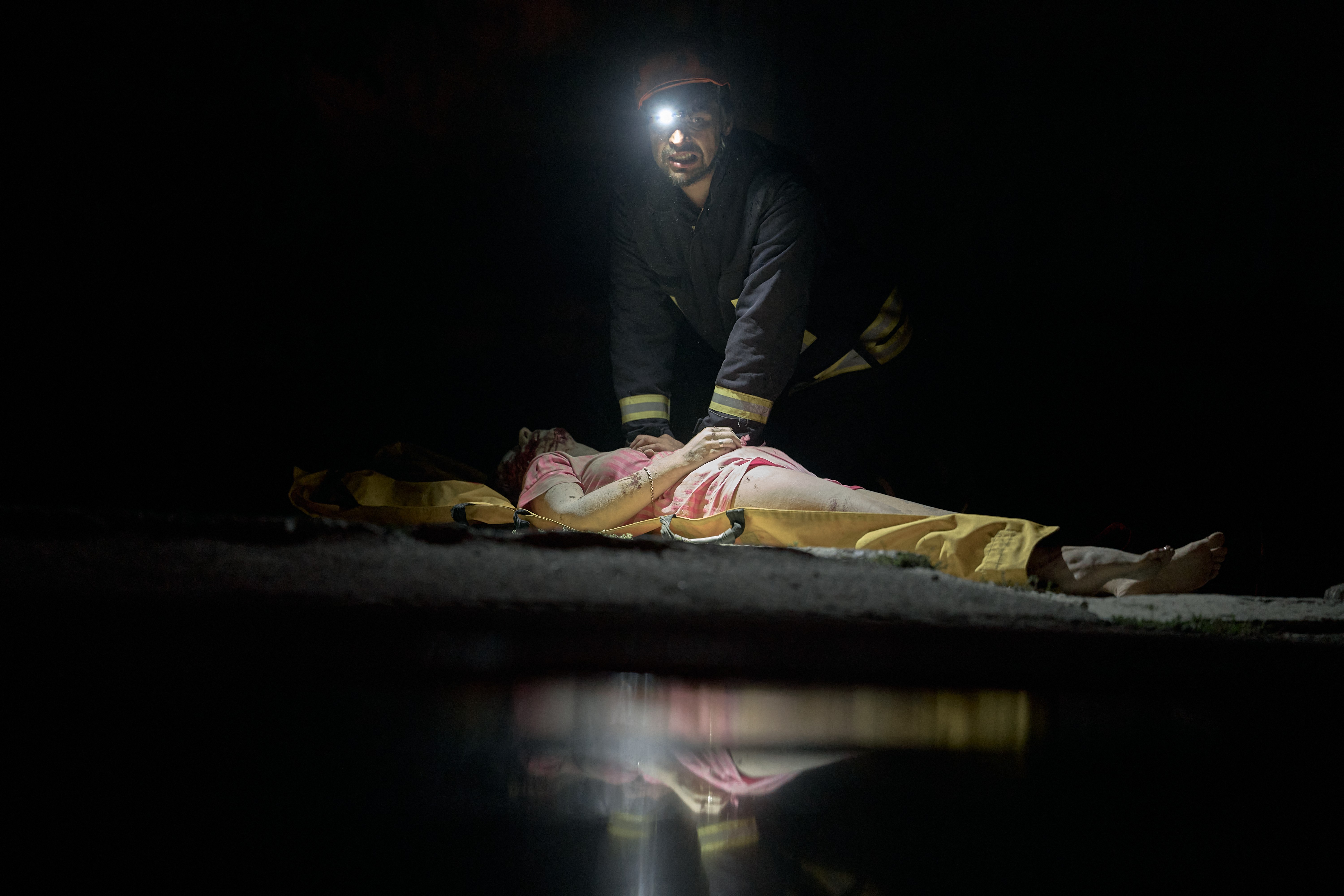 A rescue worker kneels beside an injured person on the ground, outside a damaged building at night.