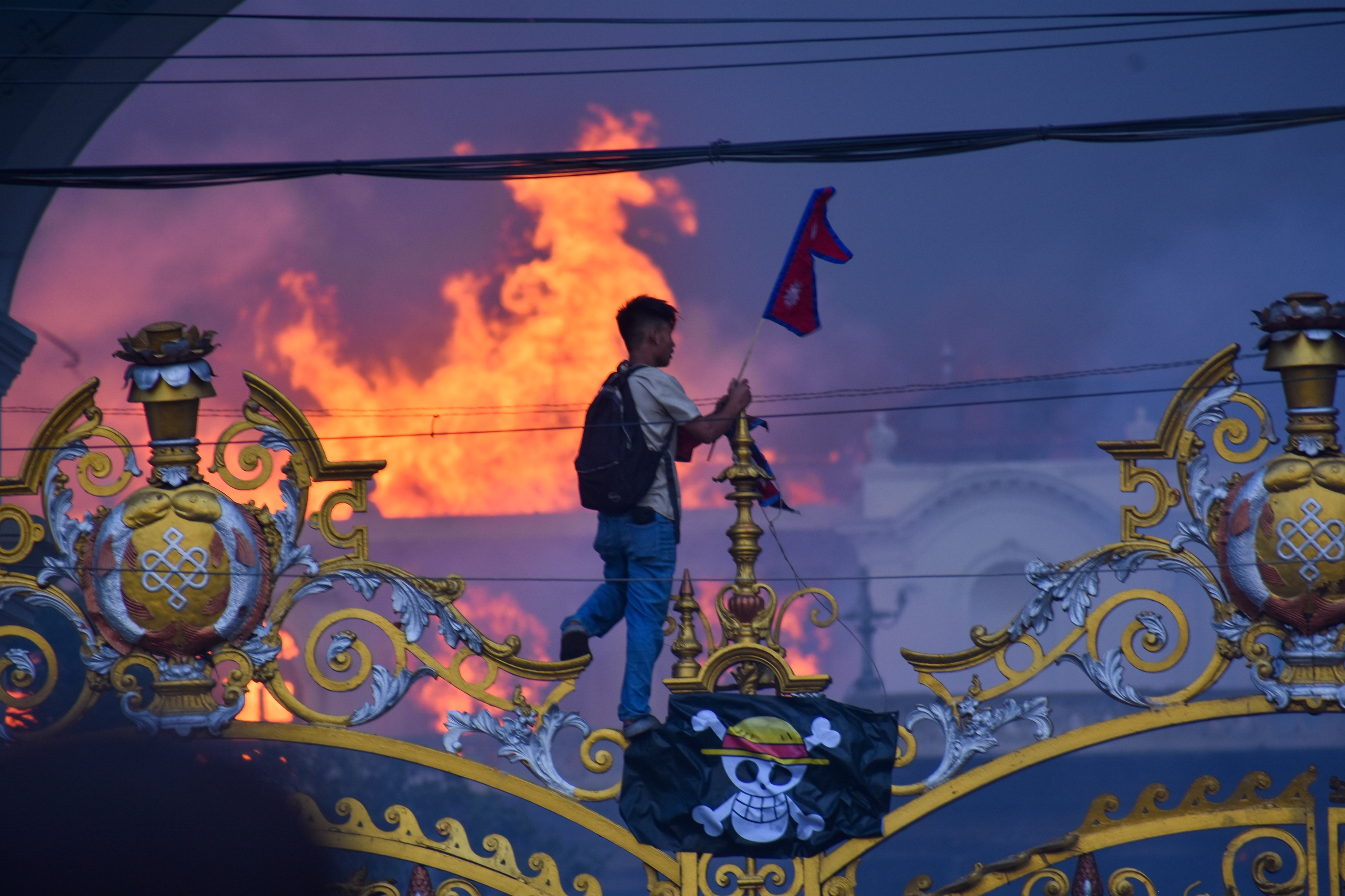 A protester carrying a Nepalese flag hangs a pirate flag on an ornate gate, as smoke and flames rise from a palace building in the background.