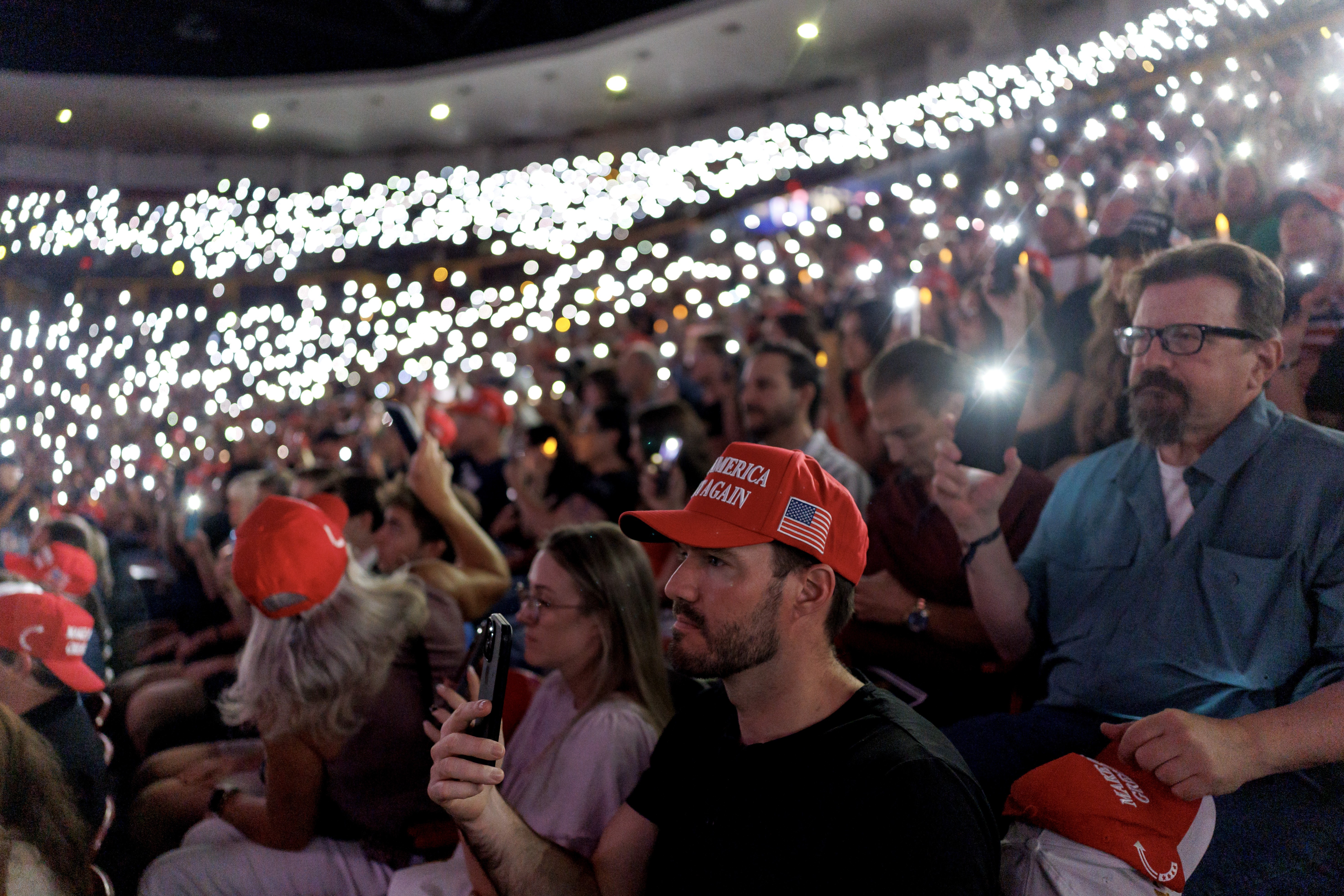 Many people sit in stadium seating, most holding up their phones with the flashlight on.