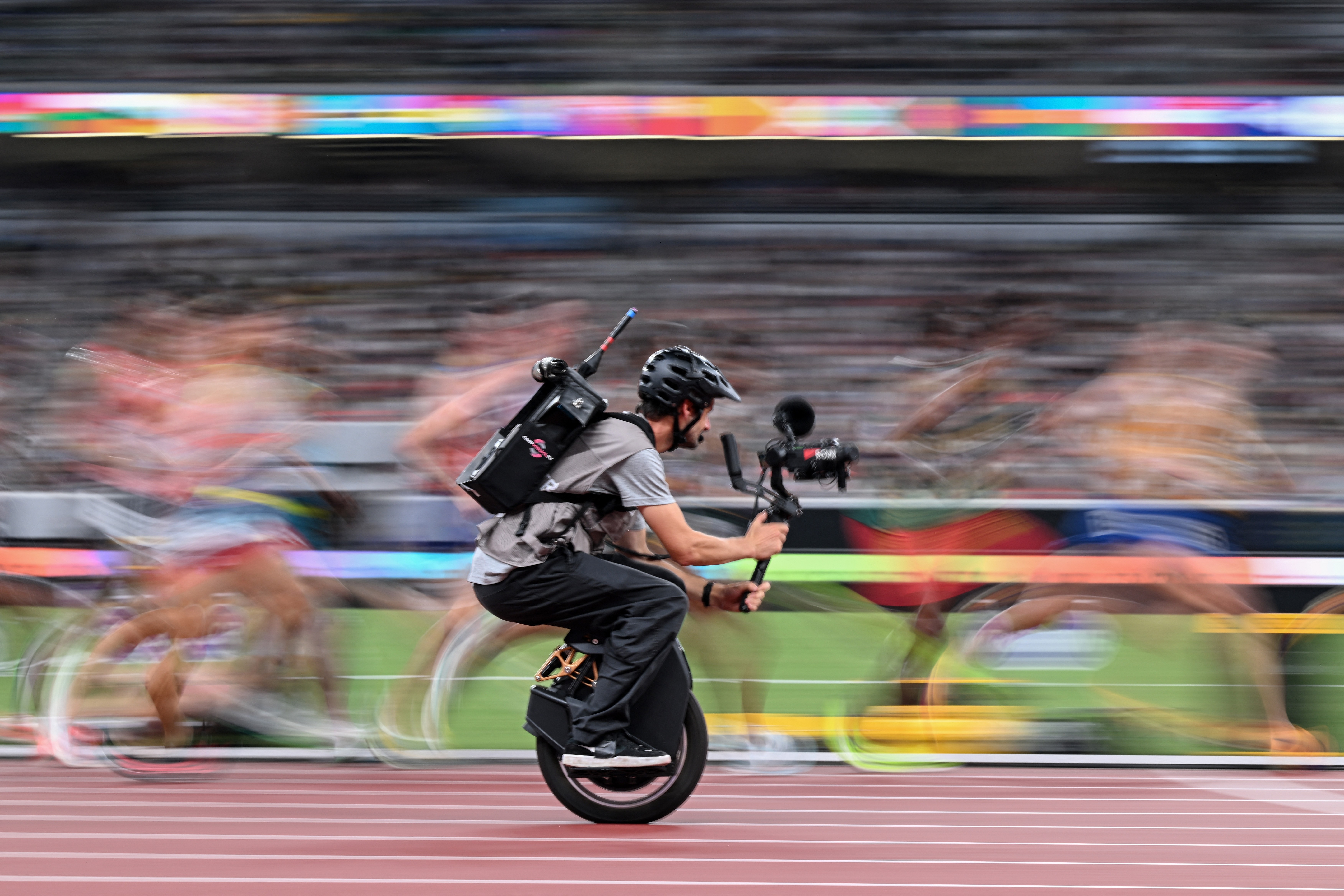 A cameraman on a unicycle films athletes running a race on a track. The runners appear blurry due to their motion.