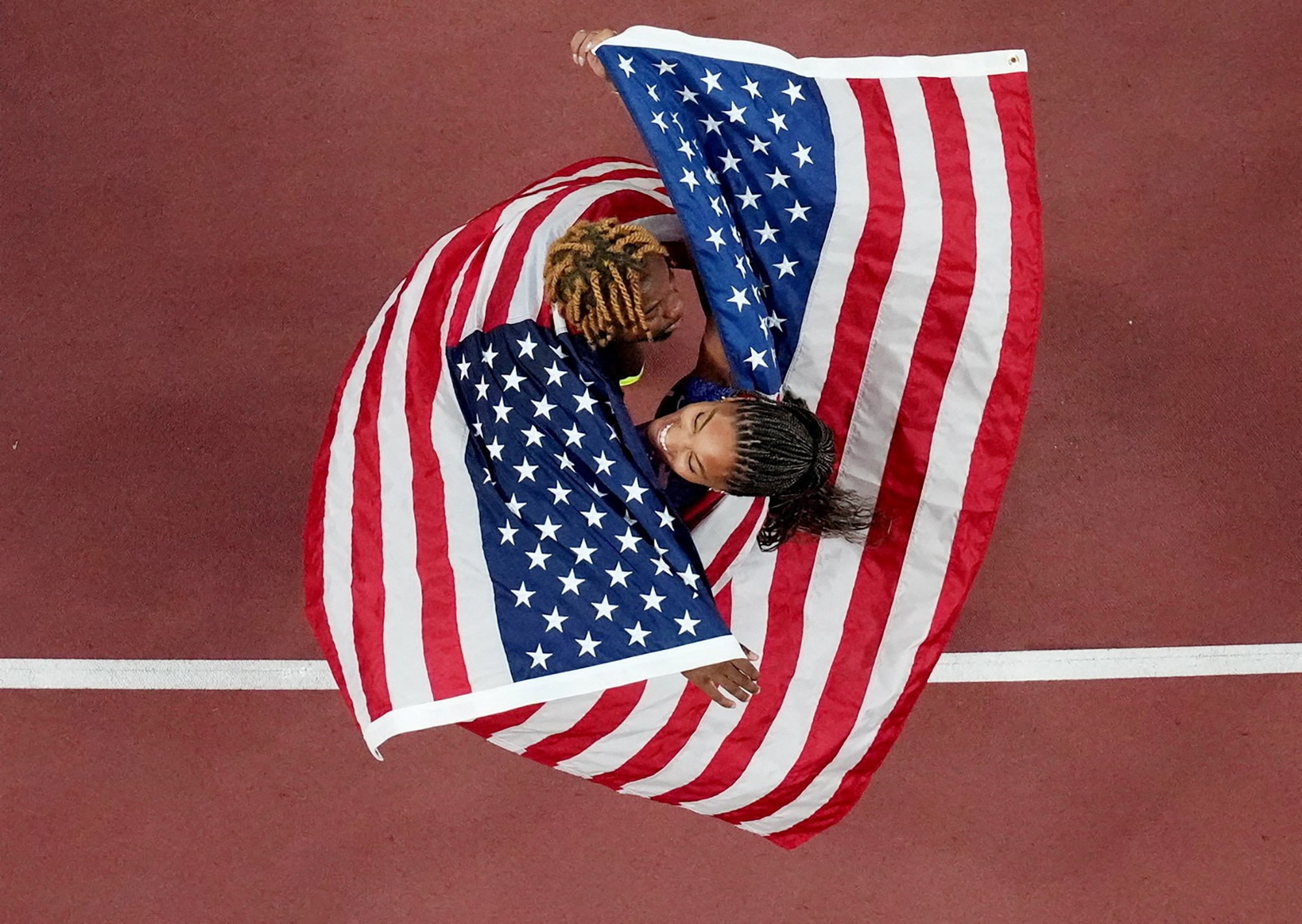 An elevated view, looking down at two athletes celebrating on a track, both wearing American flags draped around their shoulders.