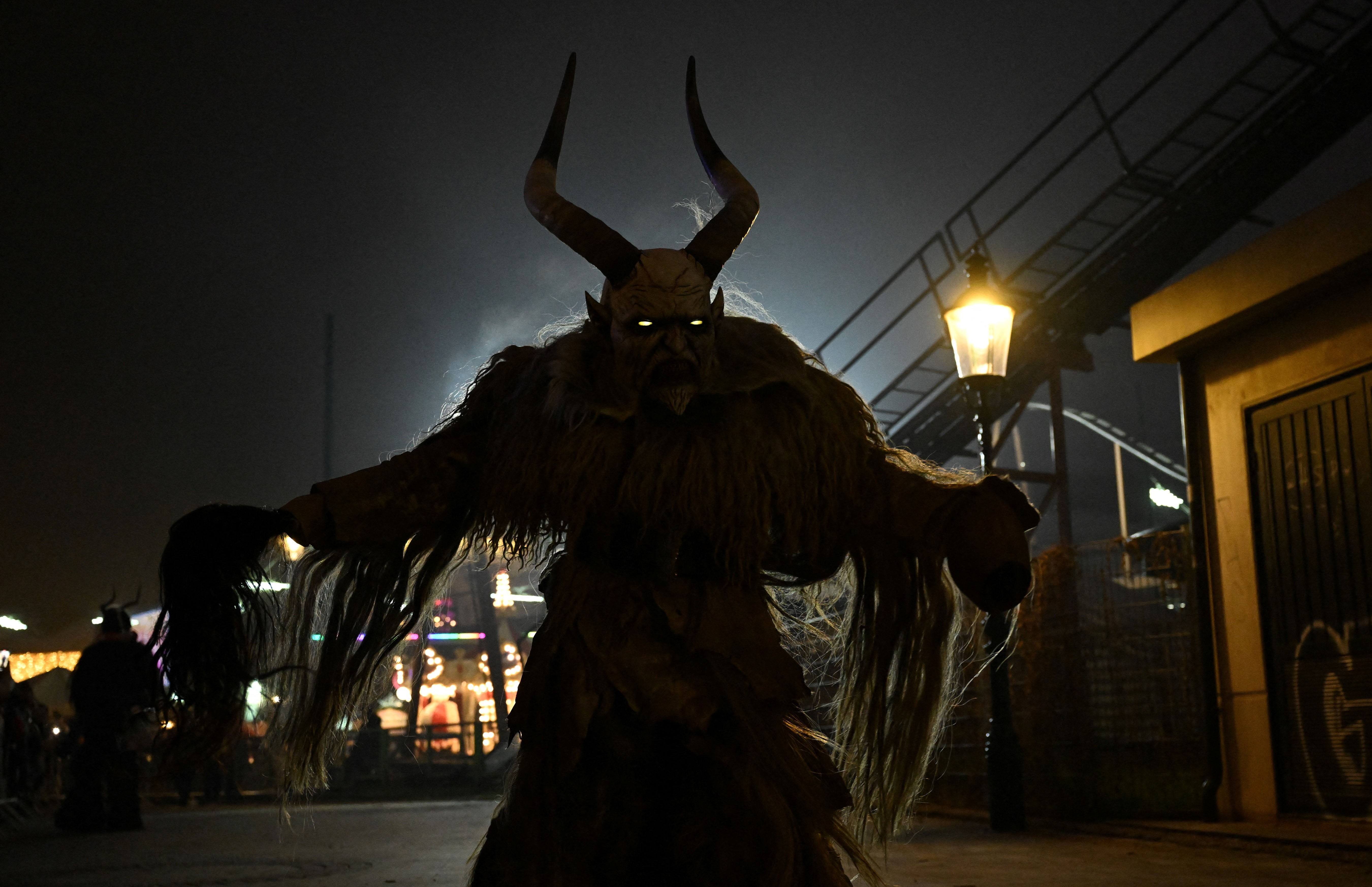 A person wearing a frightening demon mask and costume, dressed as 'Krampus,' seen at night, silhouetted by a light