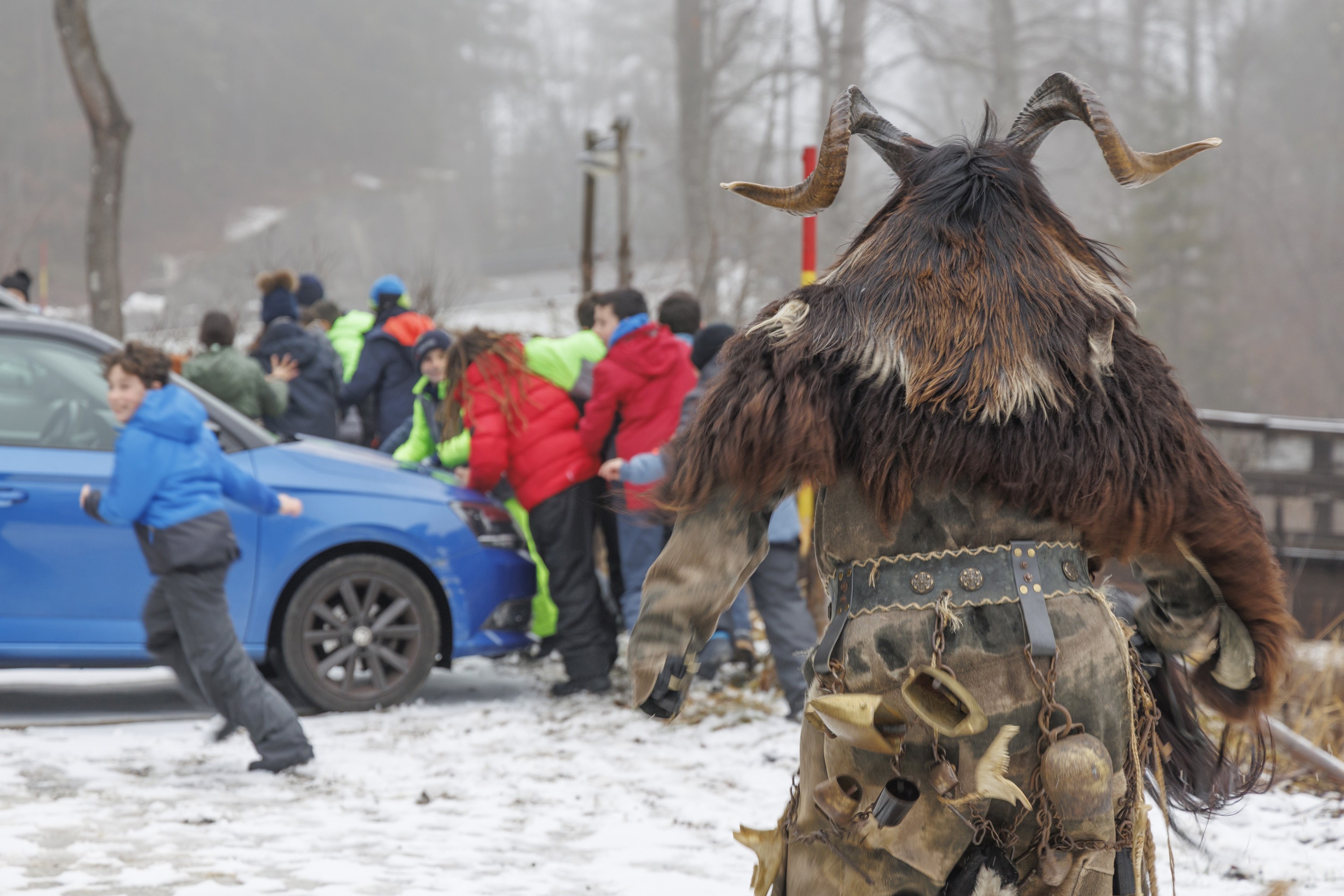 Children in a parking lot playfully run from a person wearing a scary Krampus costume.