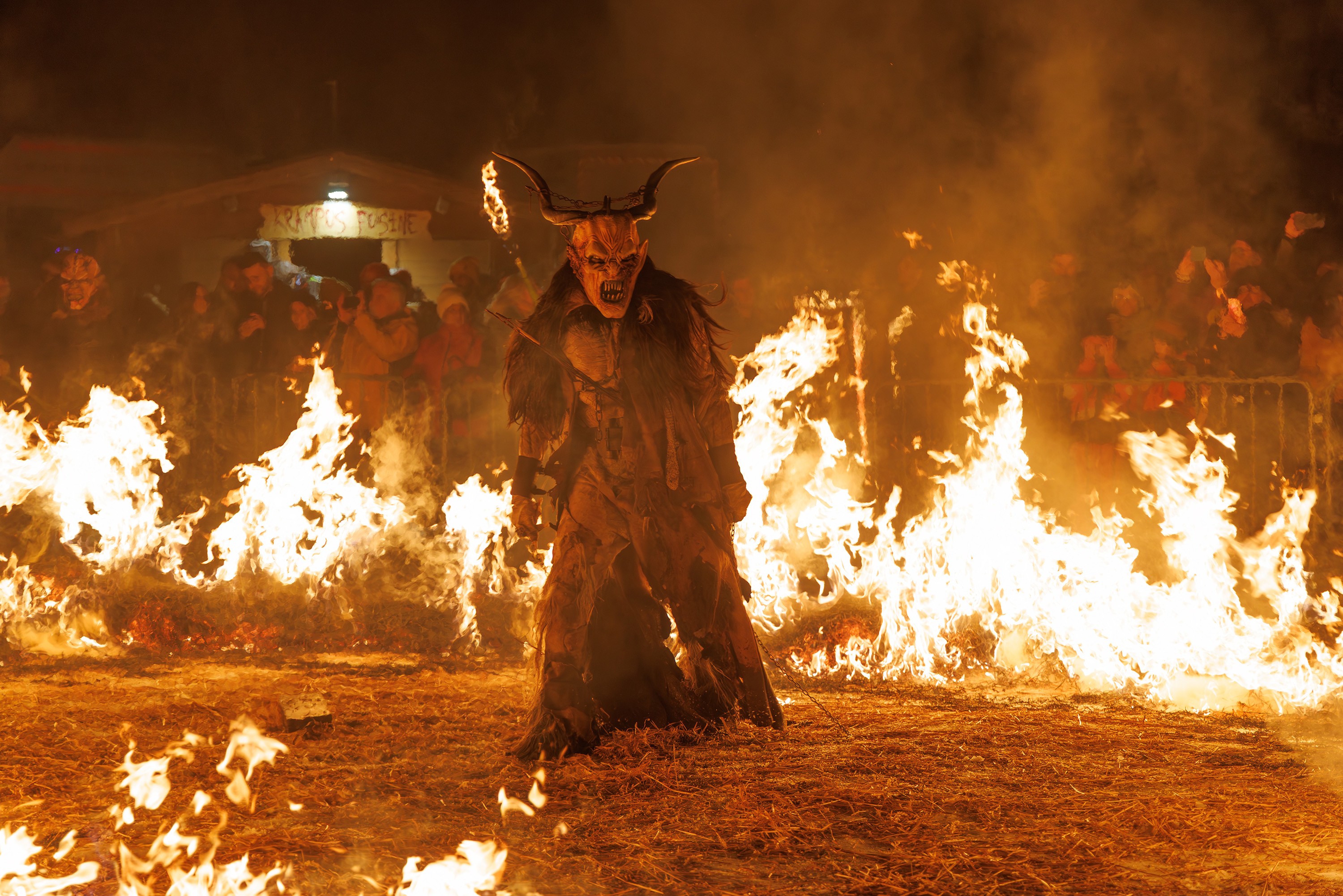 A crowd gathers to watch a person, dressed as Krampus, perform among many small fires.
