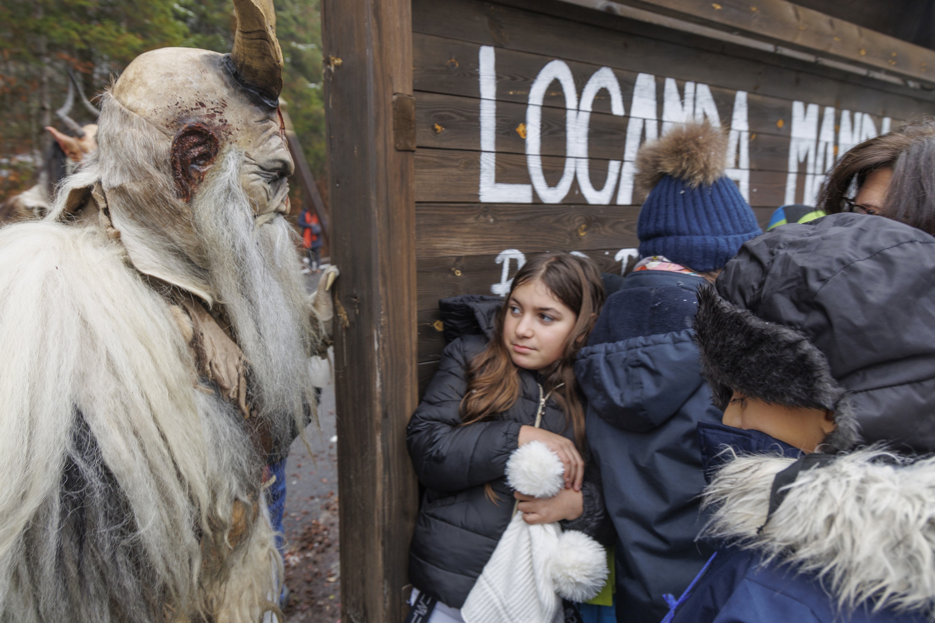 A performer dressed as Krampus approaches a group of people hiding behind a wooden sign.