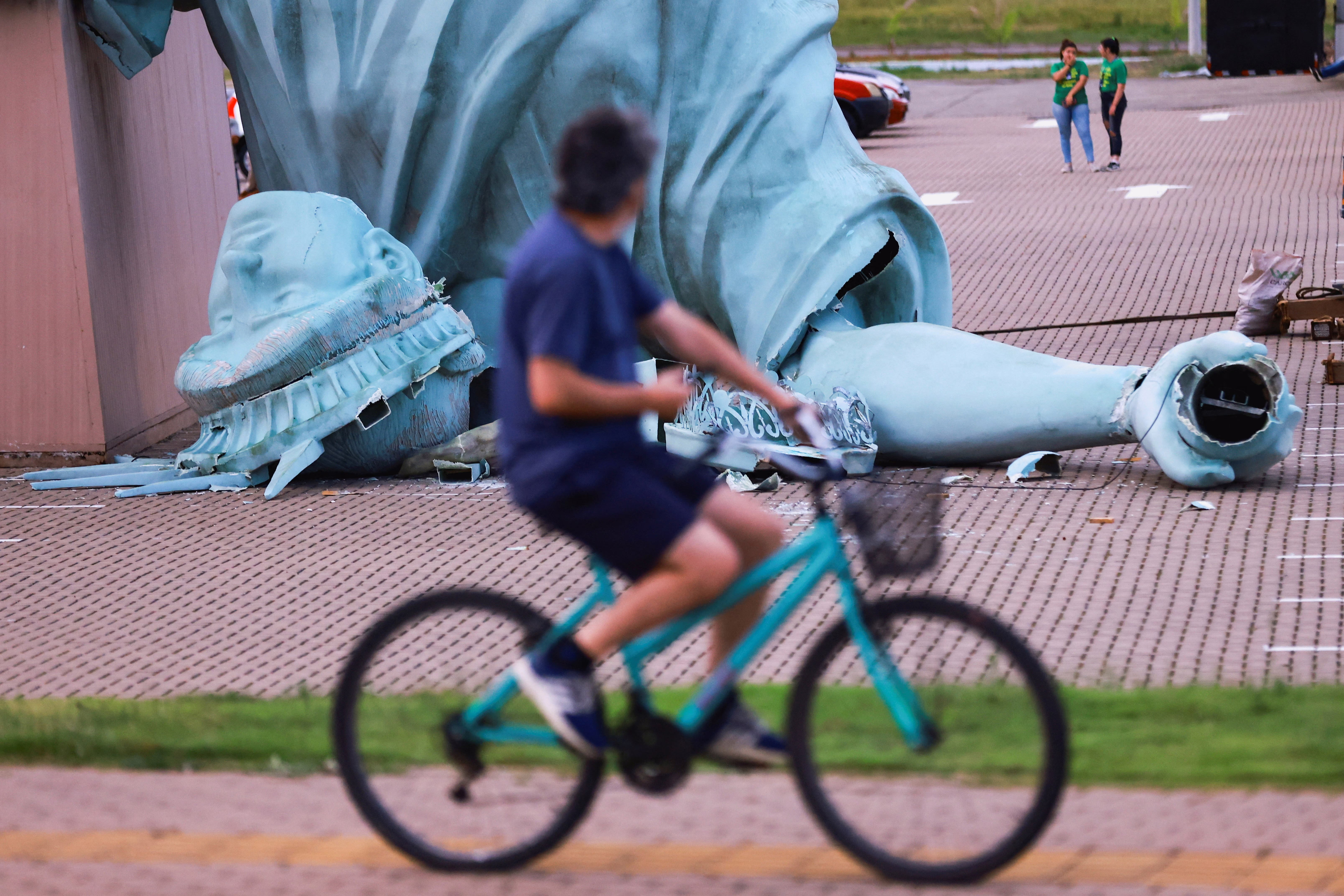 A person rides a bicycle past a toppled replica of the Statue of Liberty.