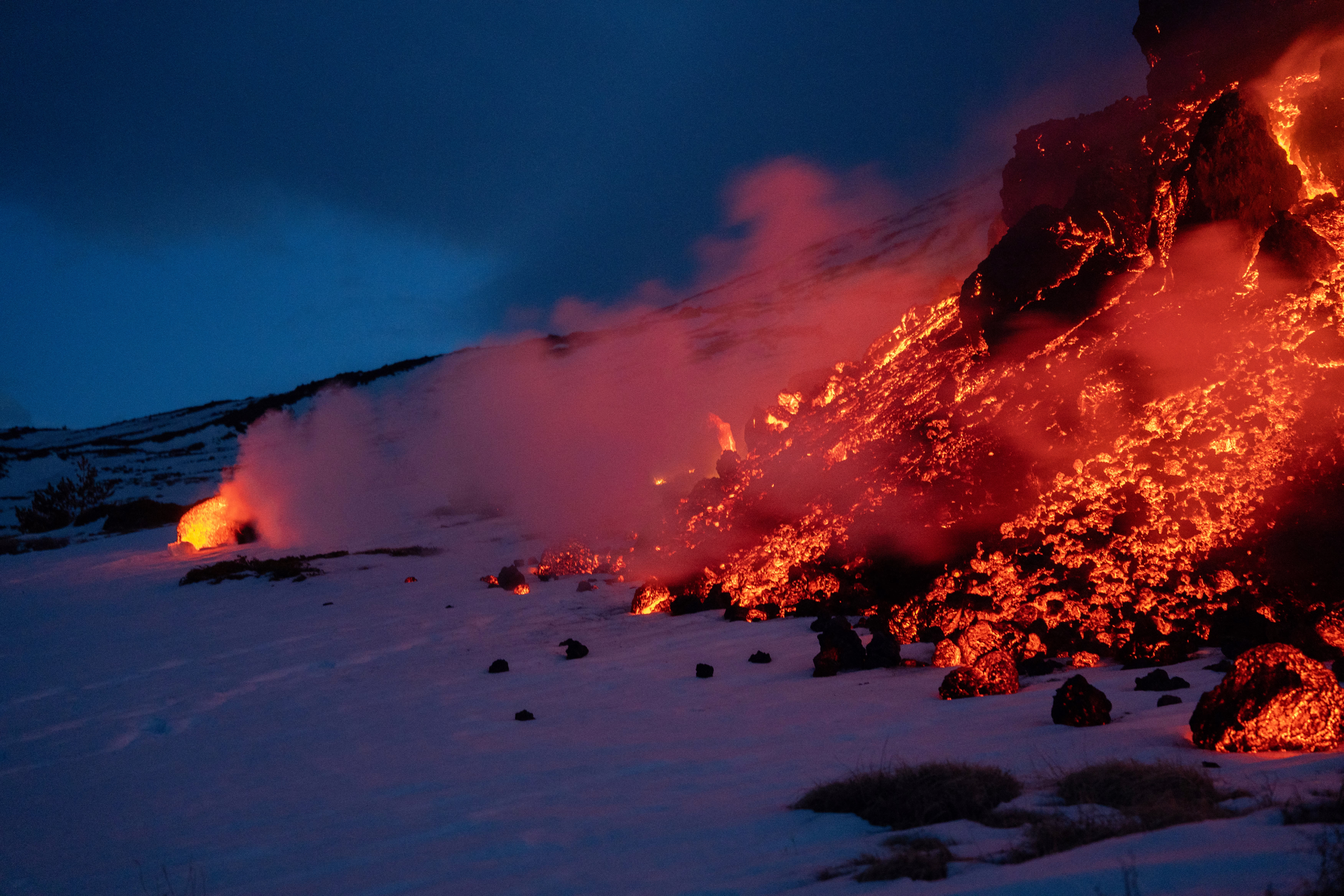 Glowing chunks of lava flow and tumble across a snow-covered slope at night.