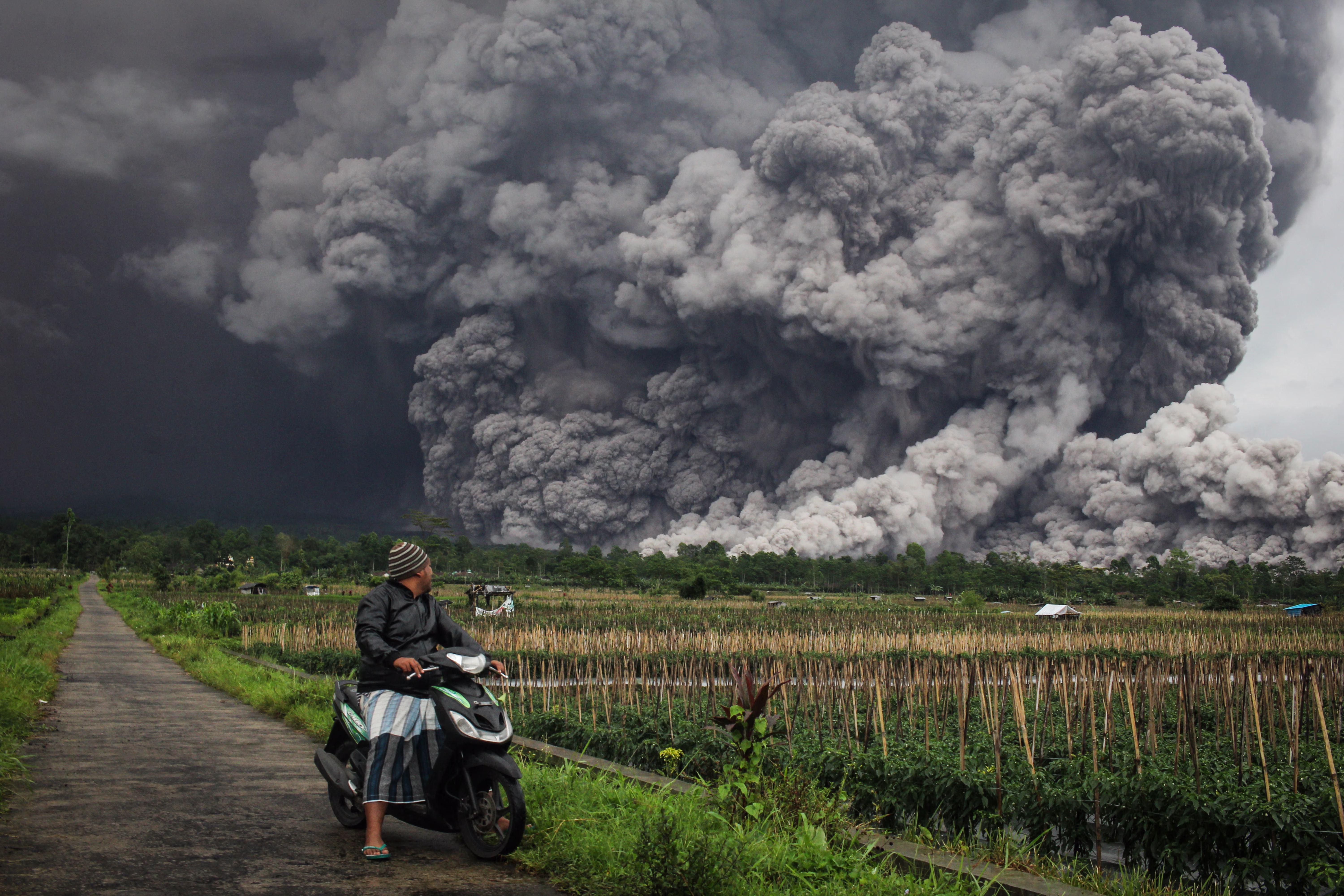 A man on a scooter looks at a huge cloud of volcanic ash billowing across a field behind him.