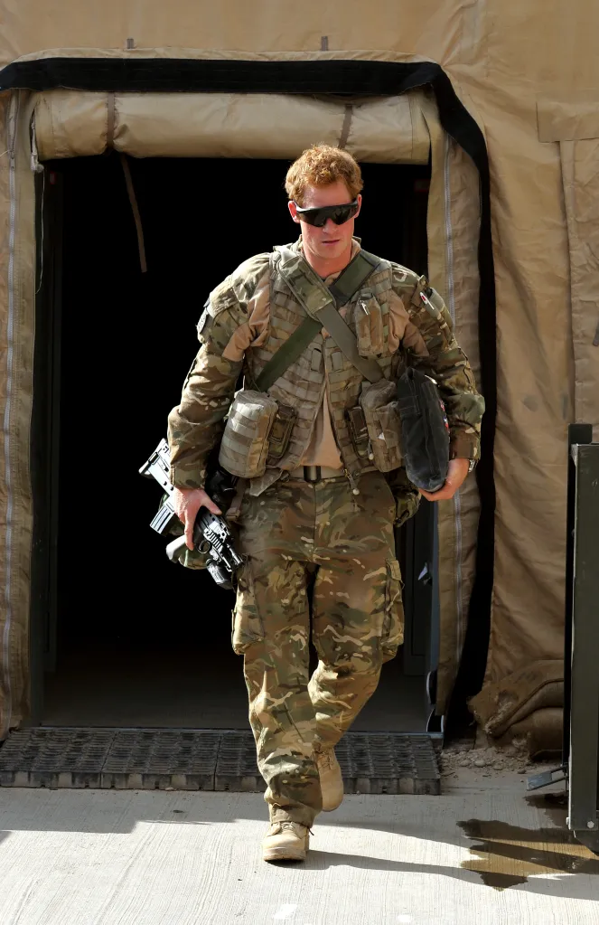 Prince Harry in camouflage uniform emerging from a tent, carrying a weapon.