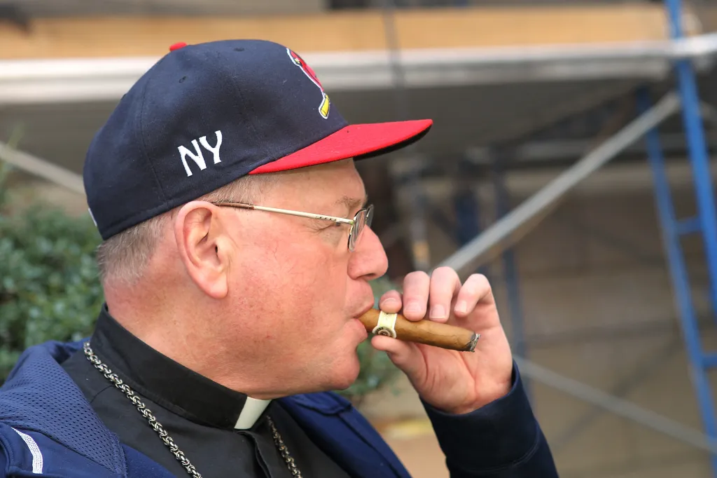 Cardinal Timothy Dolan smoking a cigar while wearing a baseball cap and glasses.