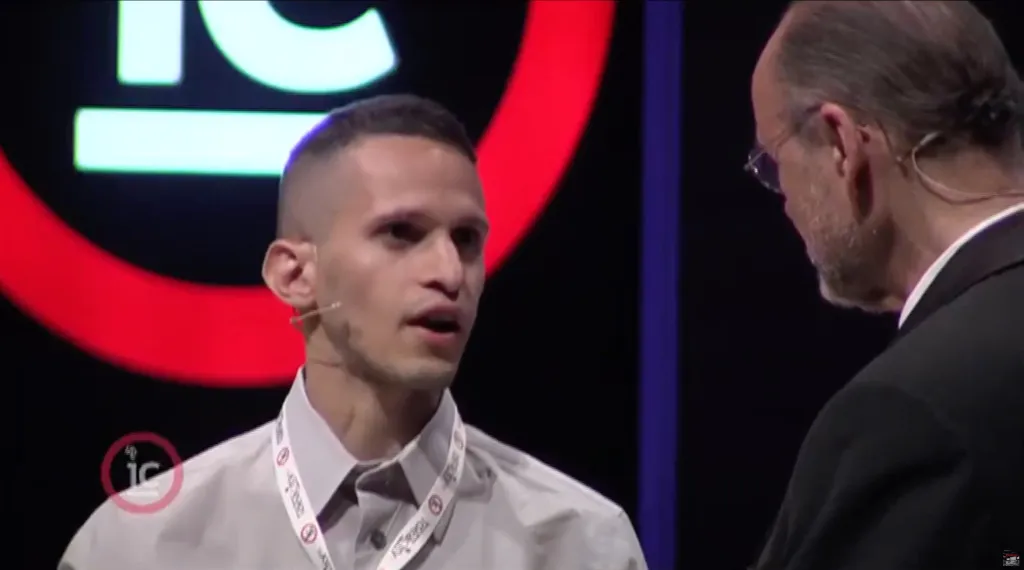 A young man speaking to an older man, both wearing business attire, on a stage with a red and black background.
