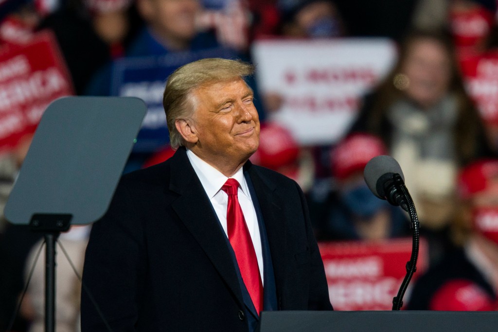 President Donald Trump smiling and looking slightly upward while wearing a black suit and a red tie at a rally.