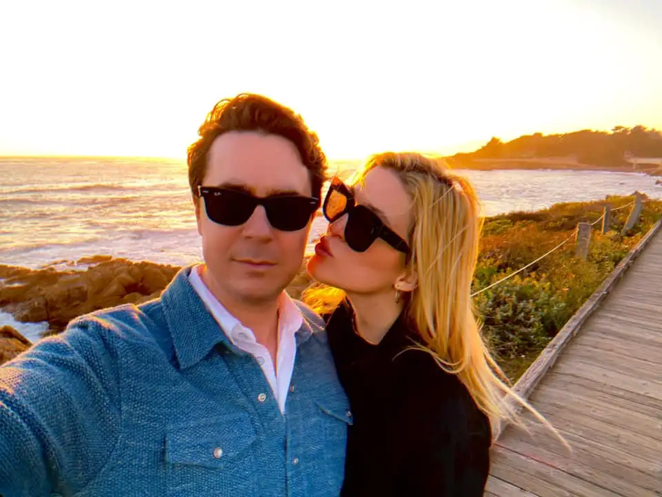 Olivia Nuzzi, in a black top and sunglasses, kissing Ryan Lizza in a blue jacket and white shirt, on the cheek with a sunset over the ocean behind them.