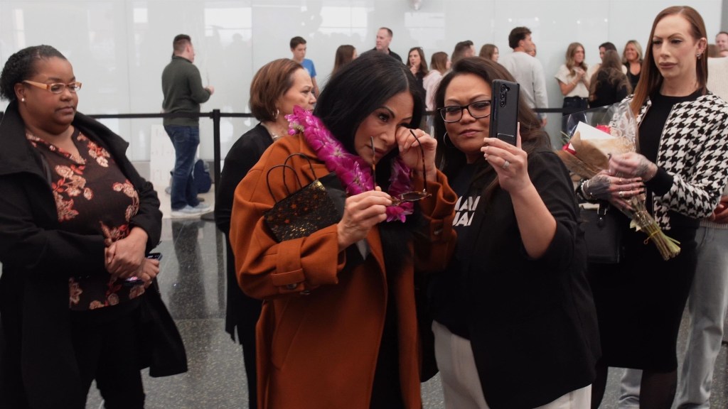 Jen Shah wiping her eye and holding sunglasses while wearing a purple lei, with another woman taking a picture with a phone.