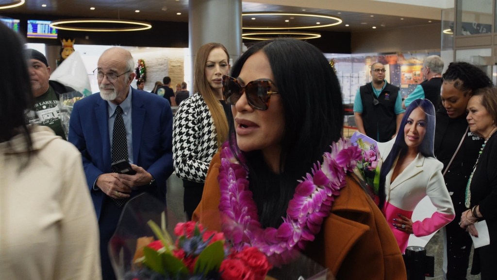 Jen Shah wearing sunglasses and a lei in an airport, surrounded by people, including a cardboard cutout of herself.