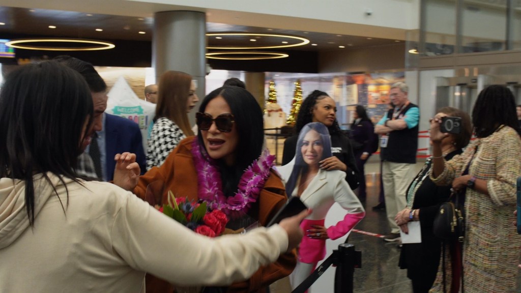 Jen Shah wearing sunglasses, a brown coat, and a purple lei, being greeted by a person holding a bouquet of red roses, with a cardboard cutout of Jen Shah in the background at an airport.